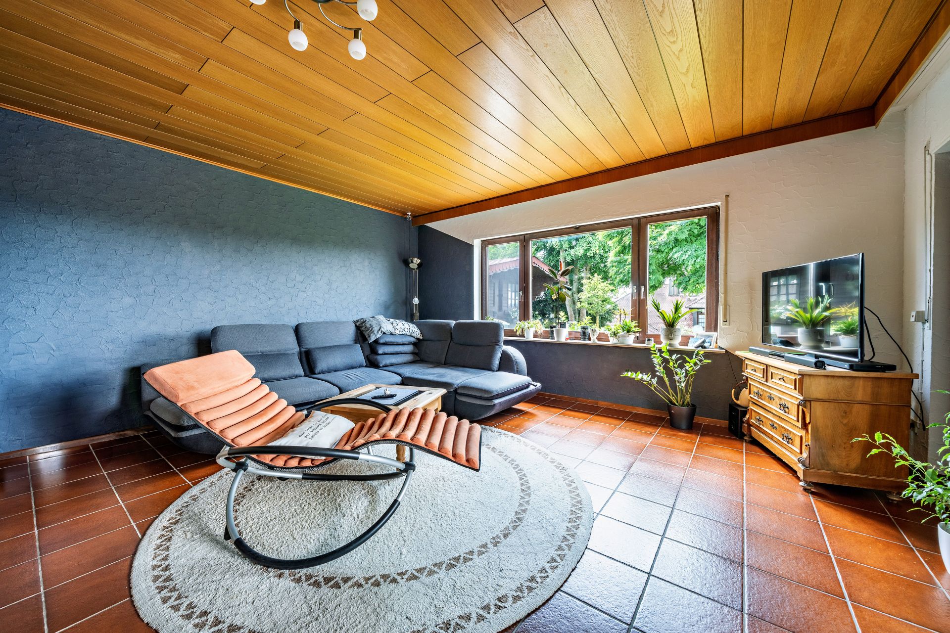 Wohnzimmer mit großer Eckcouch, Sessel, Teppich, Fernseher und vielen Pflanzen vor großen Fenstern.
