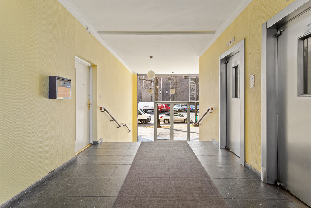 Hausflur mit gelben Wänden, Briefkasten, Eingangstür und Aufzug, Blick auf Straße mit Autos.