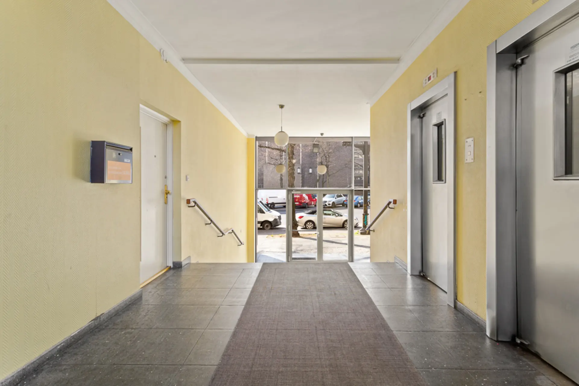 Hausflur mit gelben Wänden, Briefkasten, Eingangstür und Aufzug, Blick auf Straße mit Autos.