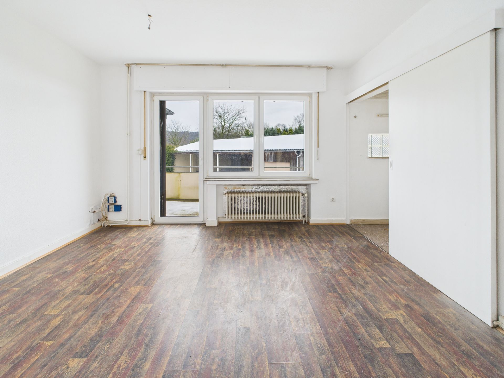Leeres Zimmer mit Holzfußboden, großen Fenstern und Zugang zu einer Terrasse.