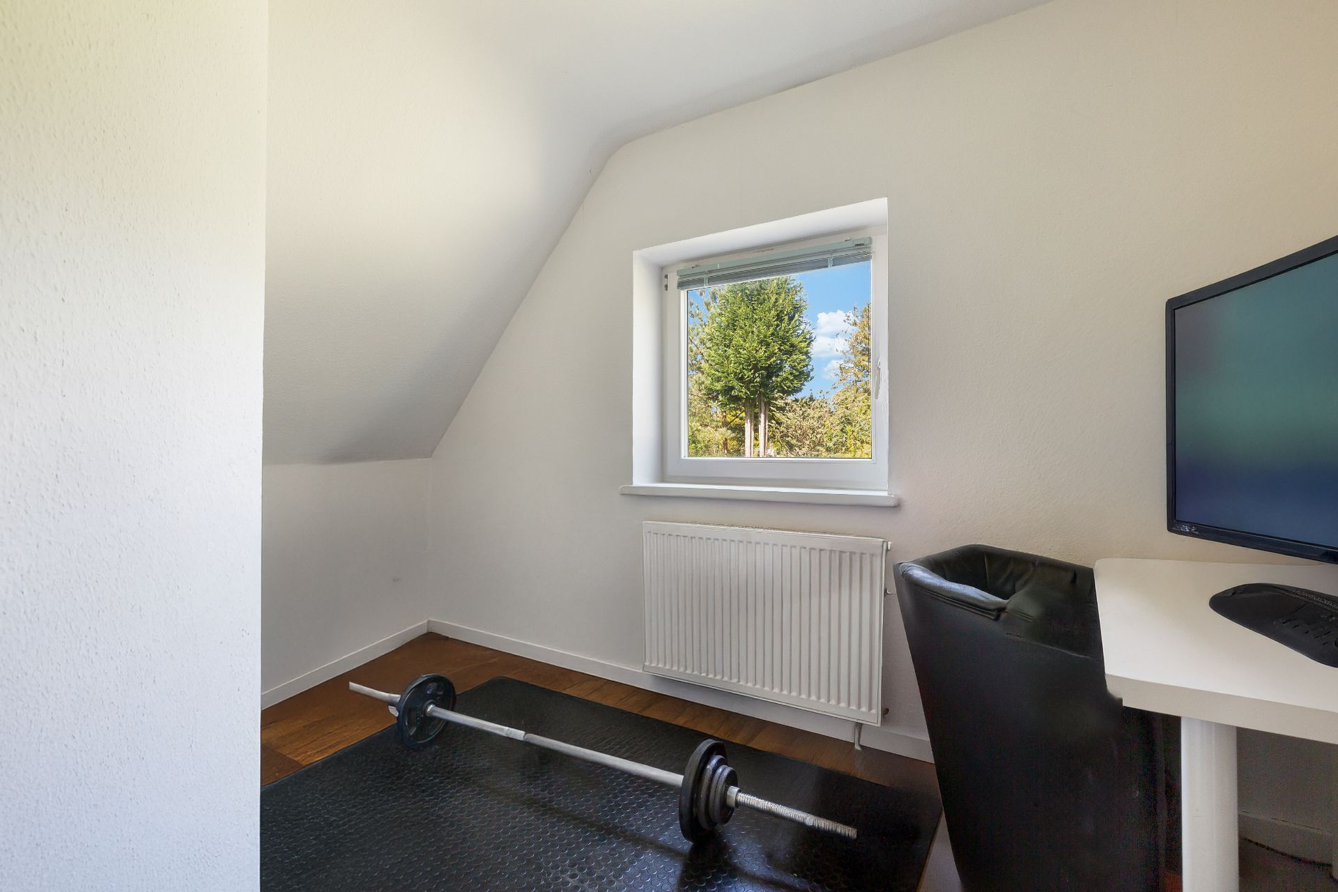 Kleines Zimmer mit Fenster, Schreibtisch, Stuhl und Hantelstange auf schwarzer Matte.
