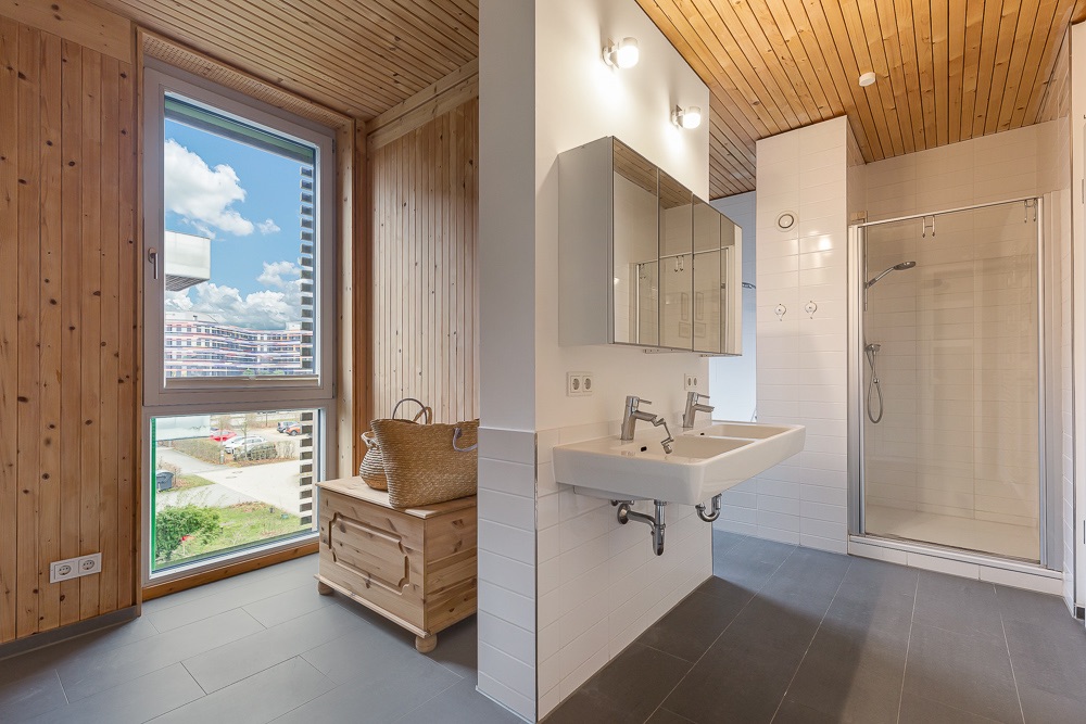 Modernes Badezimmer mit Holzwänden, Fenster, Doppelwaschbecken, Duschkabine und Holztruhe.