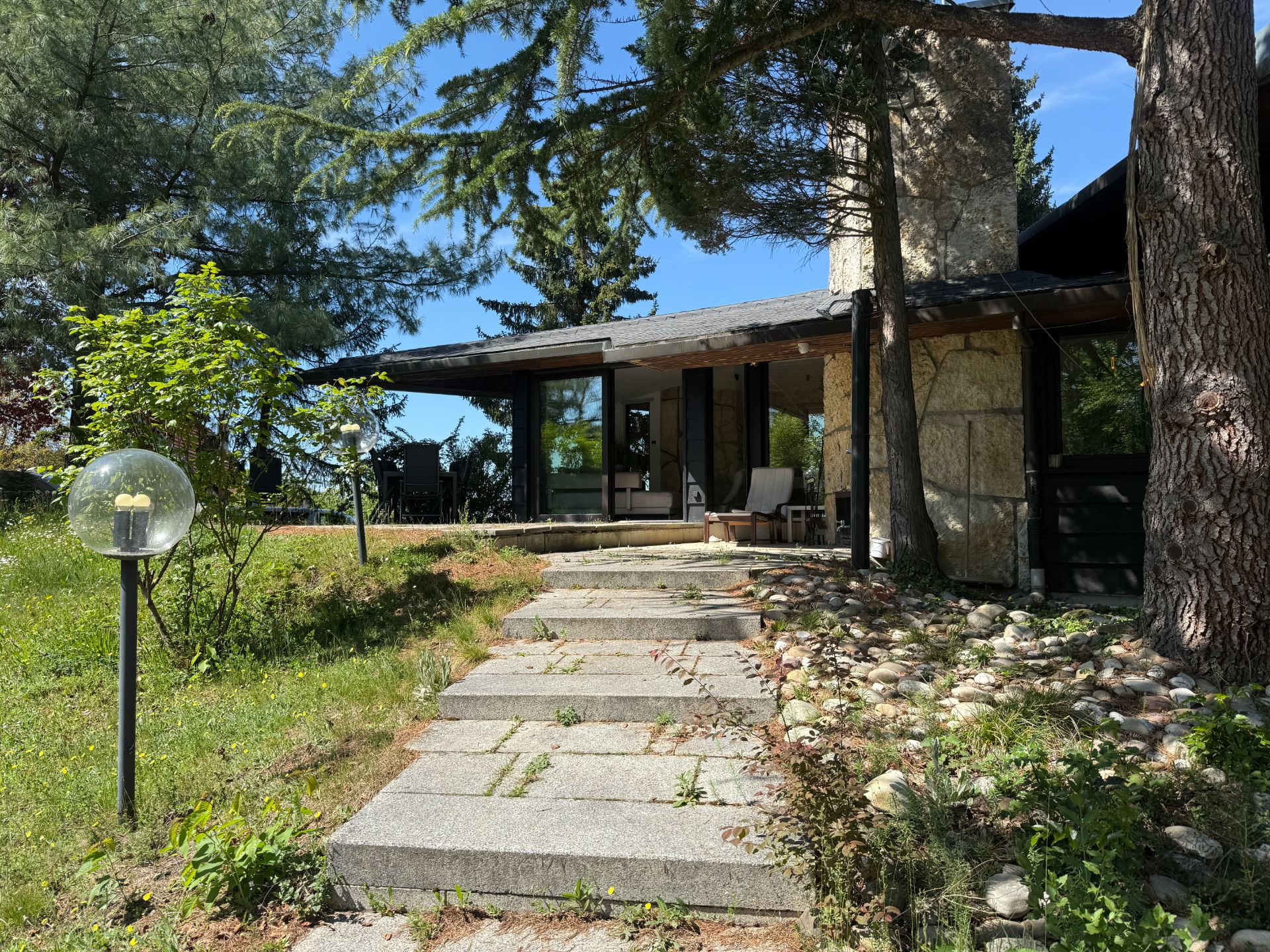 Steinweg führt zu einem Haus mit Terrasse, großen Fenstern und Bäumen im Garten.
