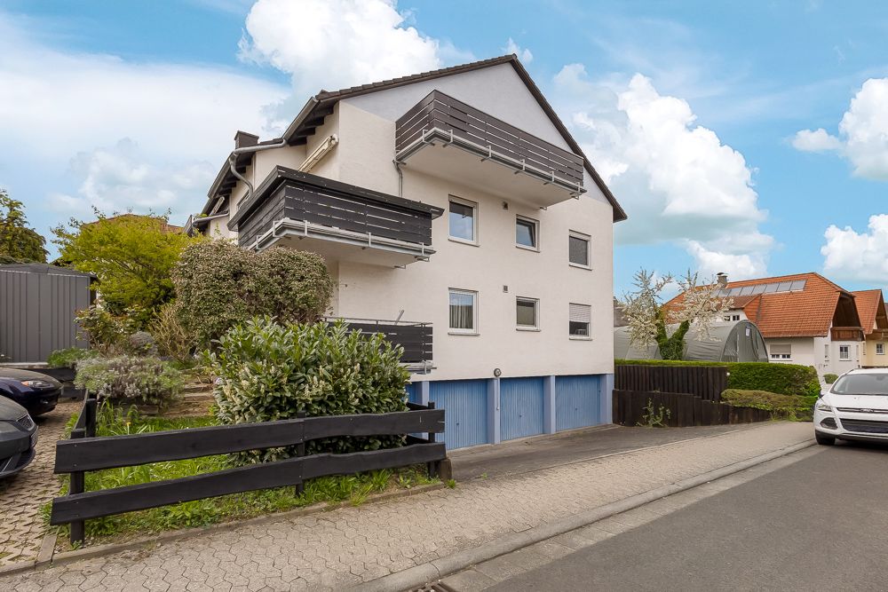 Modernes Wohnhaus mit Balkonen und blauen Garagentoren, daneben ein Garten mit Sträuchern.