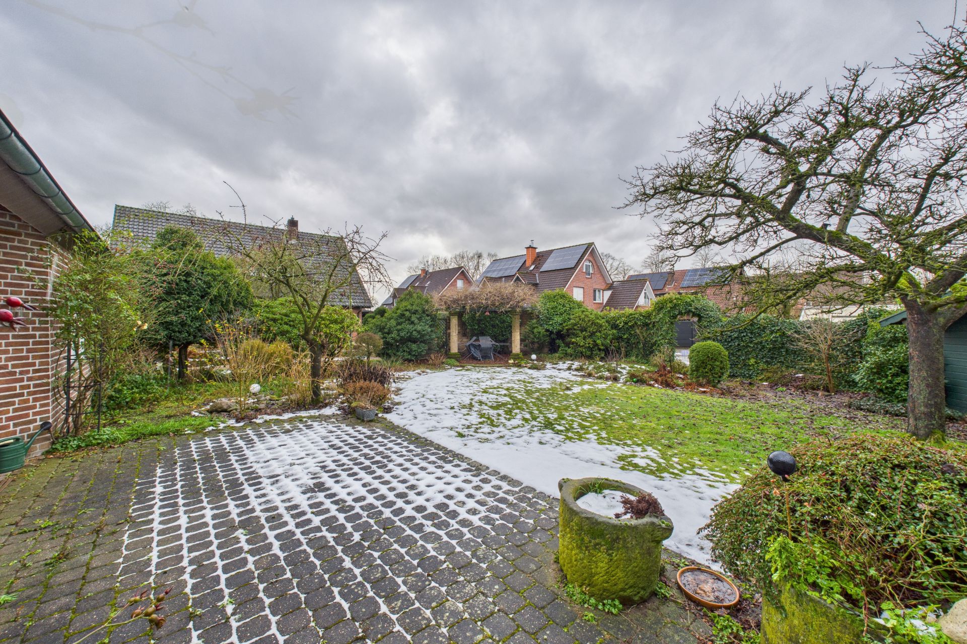 Garten mit gepflasterter Terrasse, etwas Schnee auf Rasen und Bäumen, Wohnhäuser im Hintergrund.