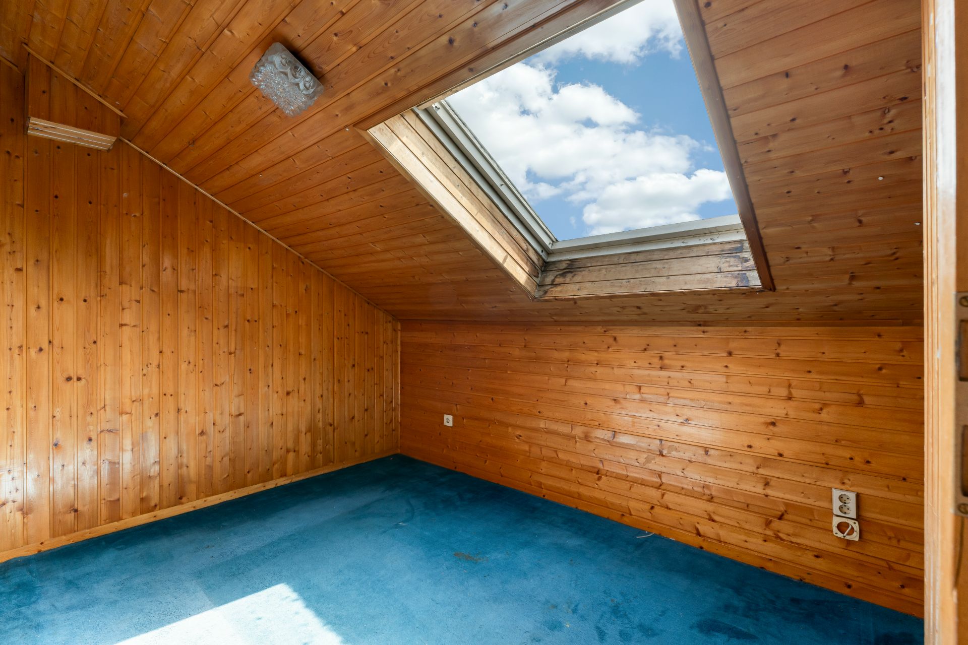 Holzvertäfelter Raum mit blauem Teppichboden und Dachflächenfenster.