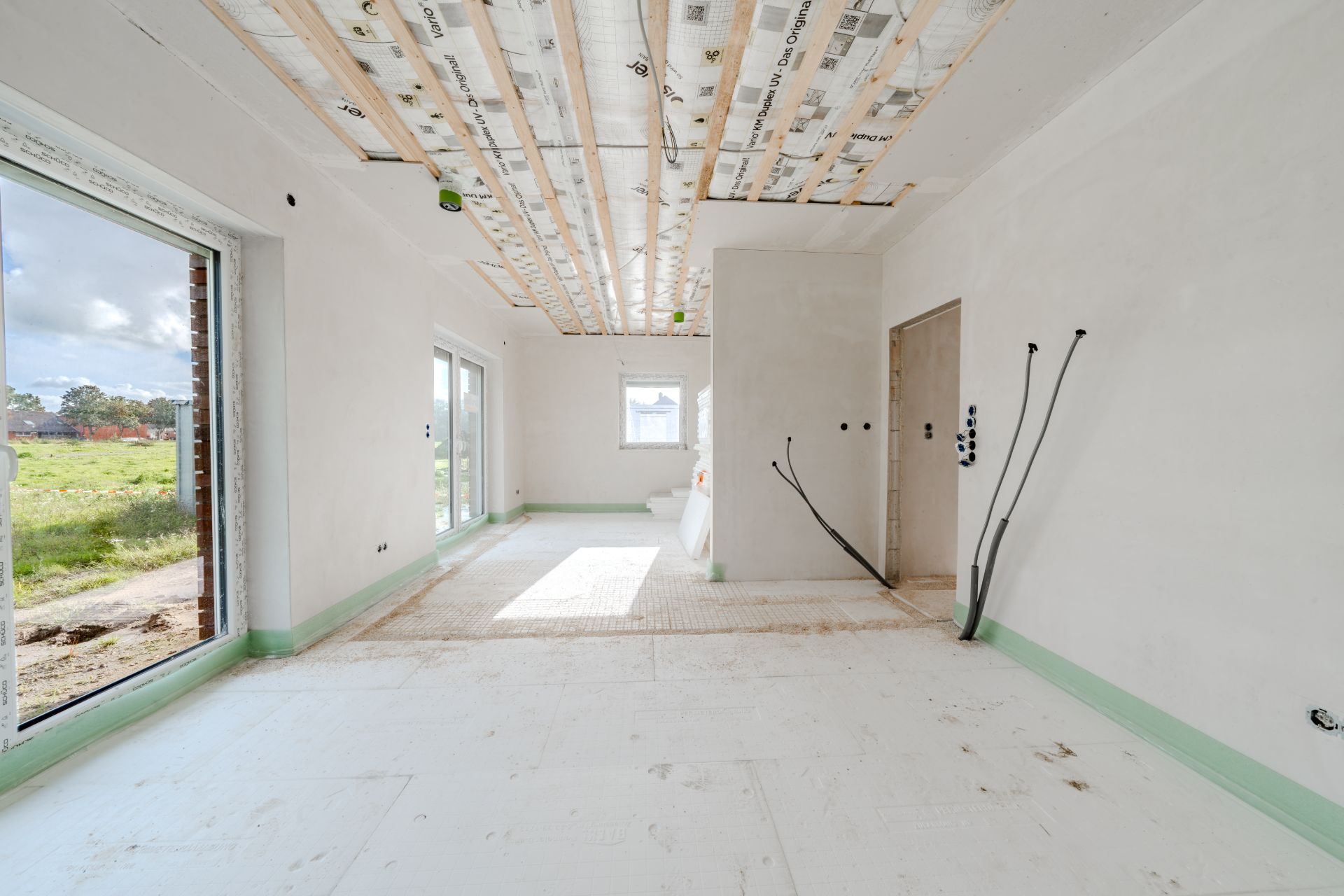 Heller Raum in einem Rohbauhaus mit großen Fenstern und offenen Kabeln an der Wand.