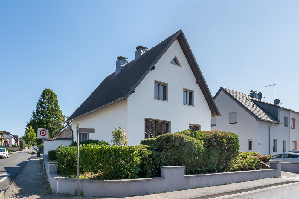 Weißes Einfamilienhaus mit dunklem Satteldach und gepflegtem Vorgarten an einer Straßenecke.