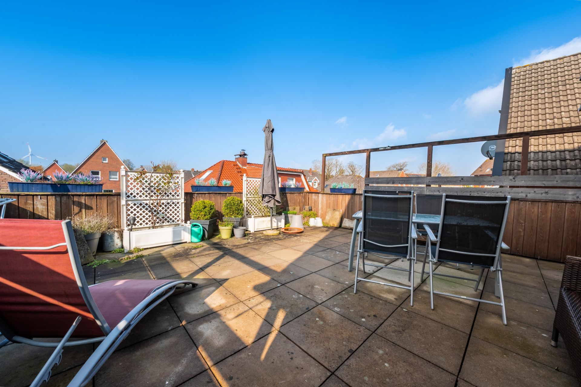 Große Terrasse mit Liegestuhl, Tisch, Stühlen, Pflanzen und Sonnenschirm, Blick auf Nachbarhäuser.