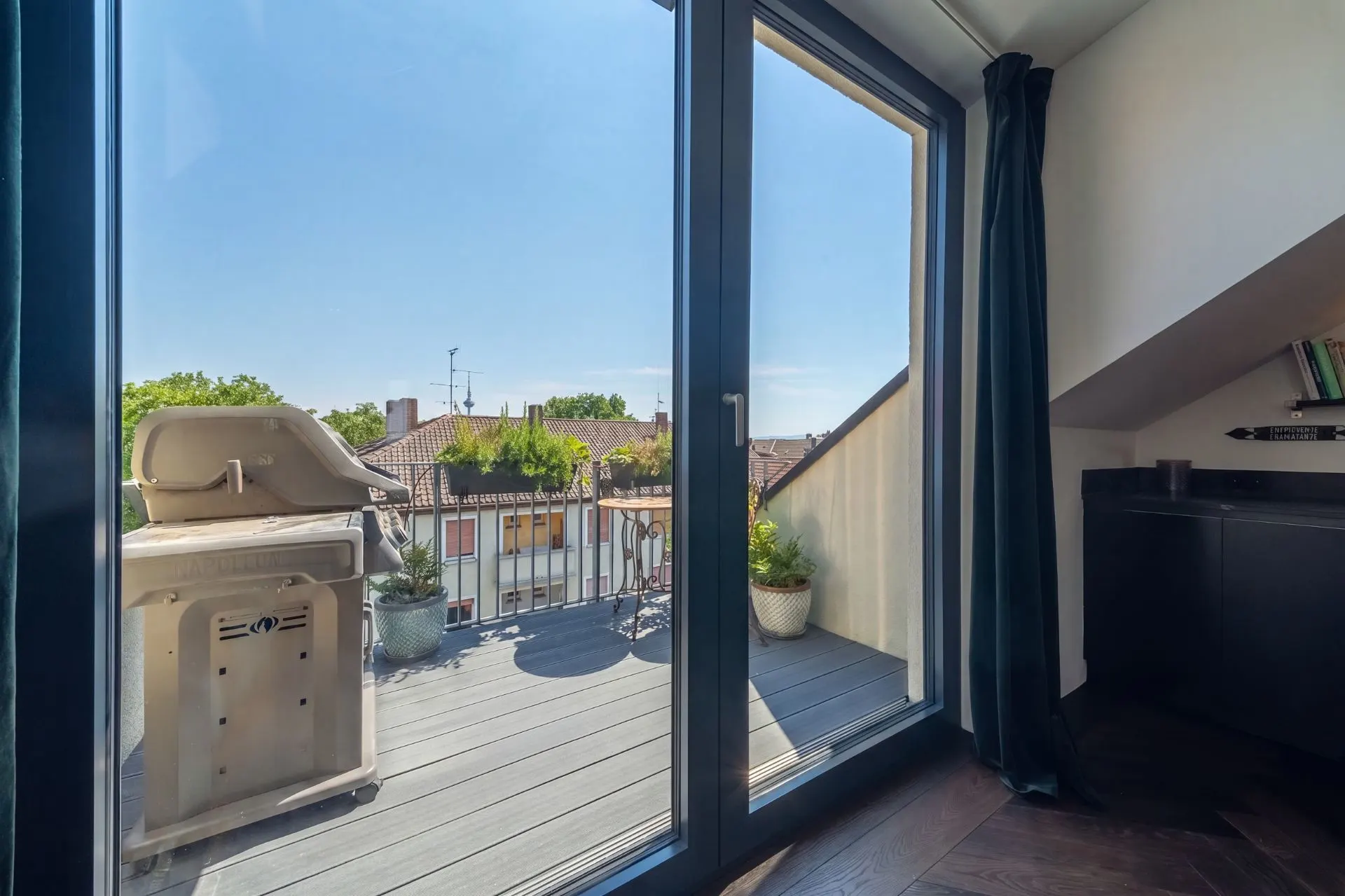Balkon mit Grill, Pflanzentöpfen und Blick auf Dächer, von hellem Raum aus fotografiert.