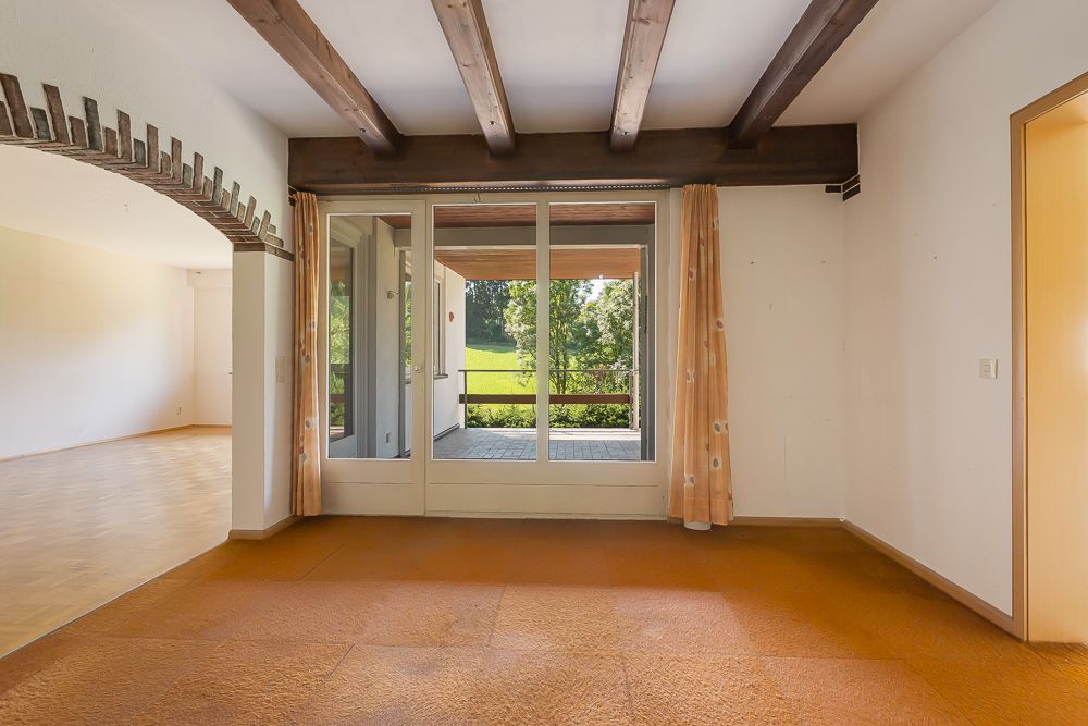 Wohnzimmer mit Glastür zur Terrasse, Holzbalken an der Decke und orangefarbenem Teppichboden.