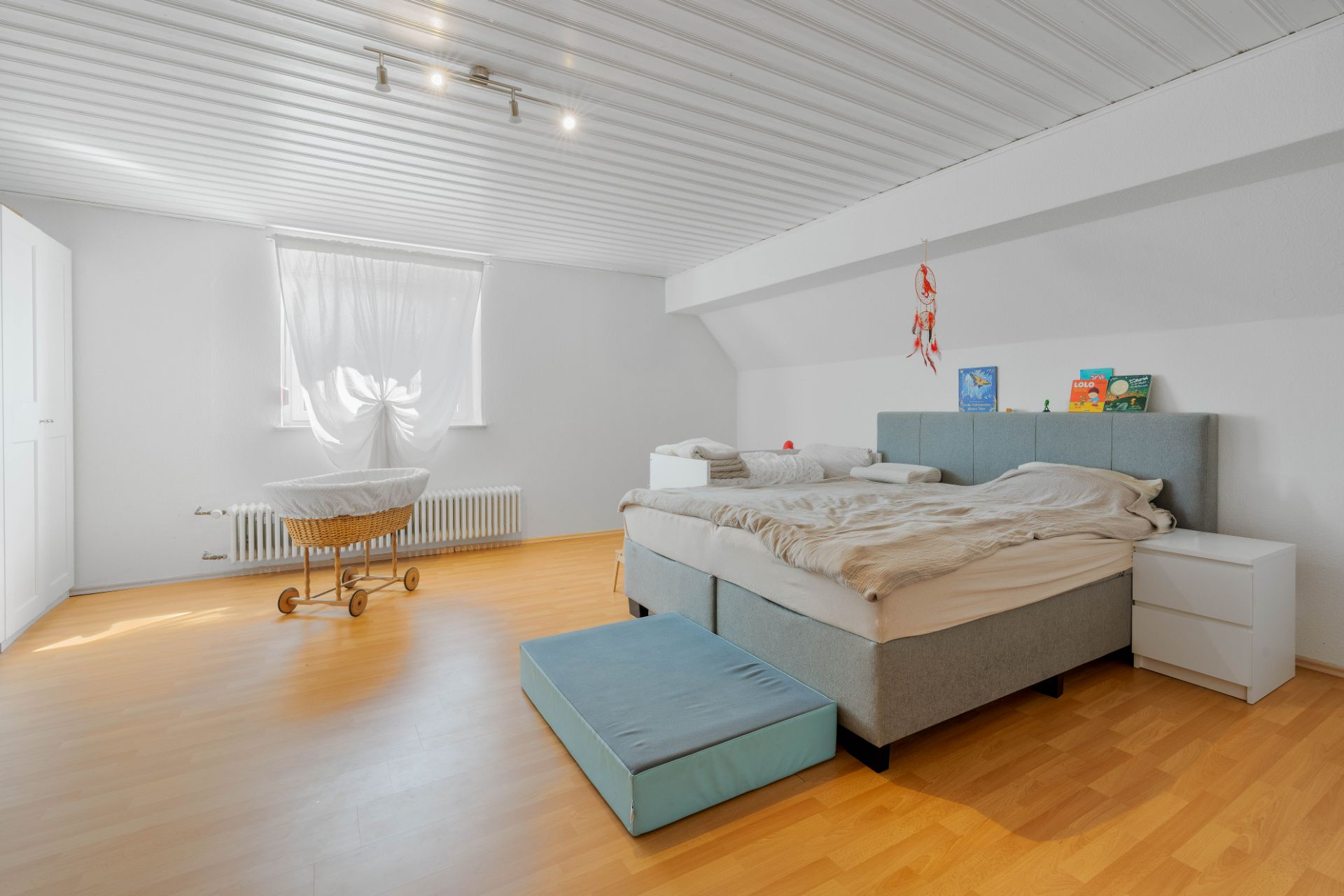 Helles Schlafzimmer mit Doppelbett, Nachttisch, Babybett und Fenster mit Vorhang.