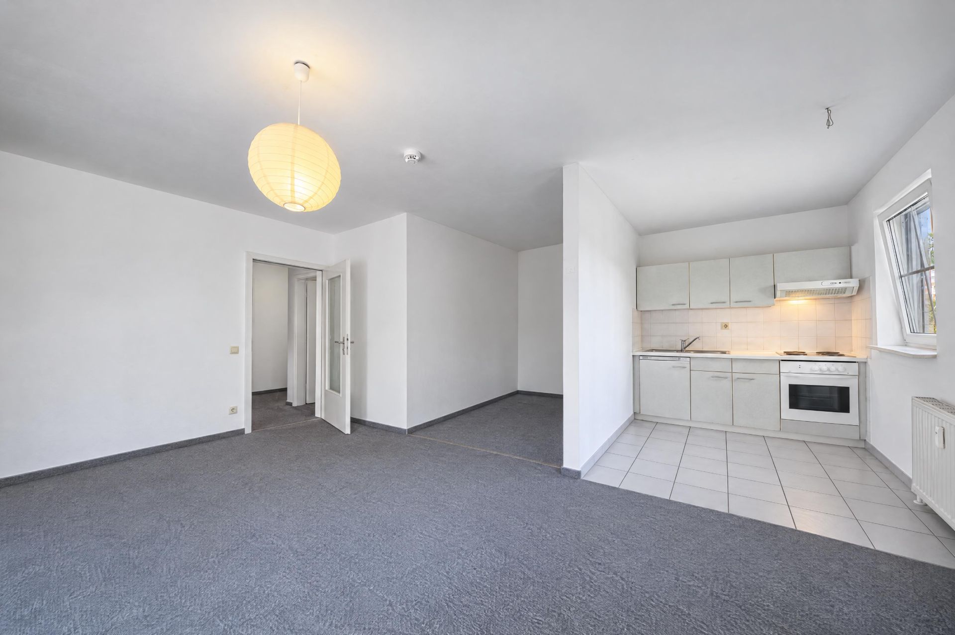 Helles Wohnzimmer mit offener Einbauküche, grauem Teppich und großer Fensterfront.