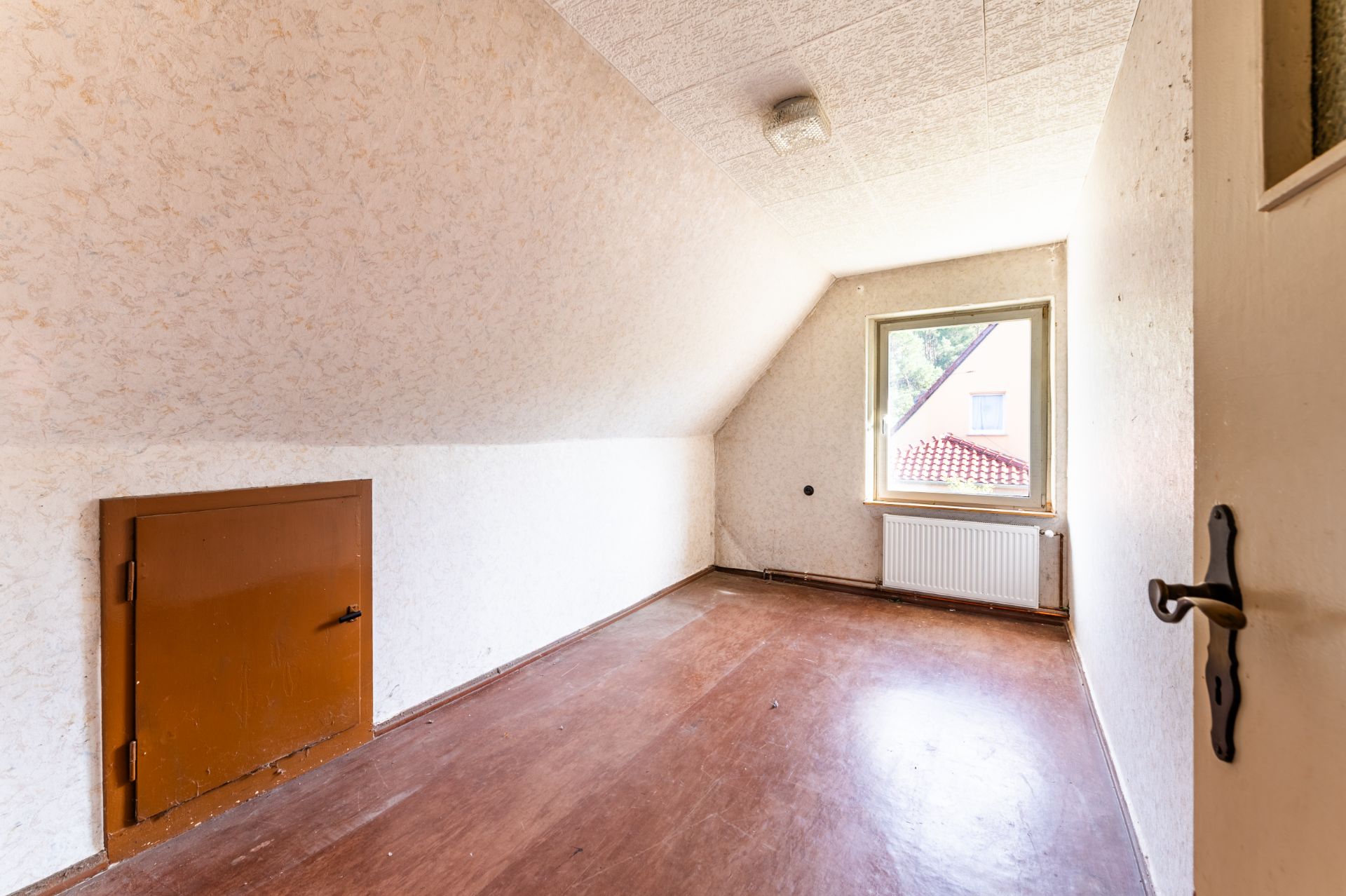 Kleiner leerer Raum mit braunem Boden, Dachschräge, Fenster und kleinem Wandtürchen.