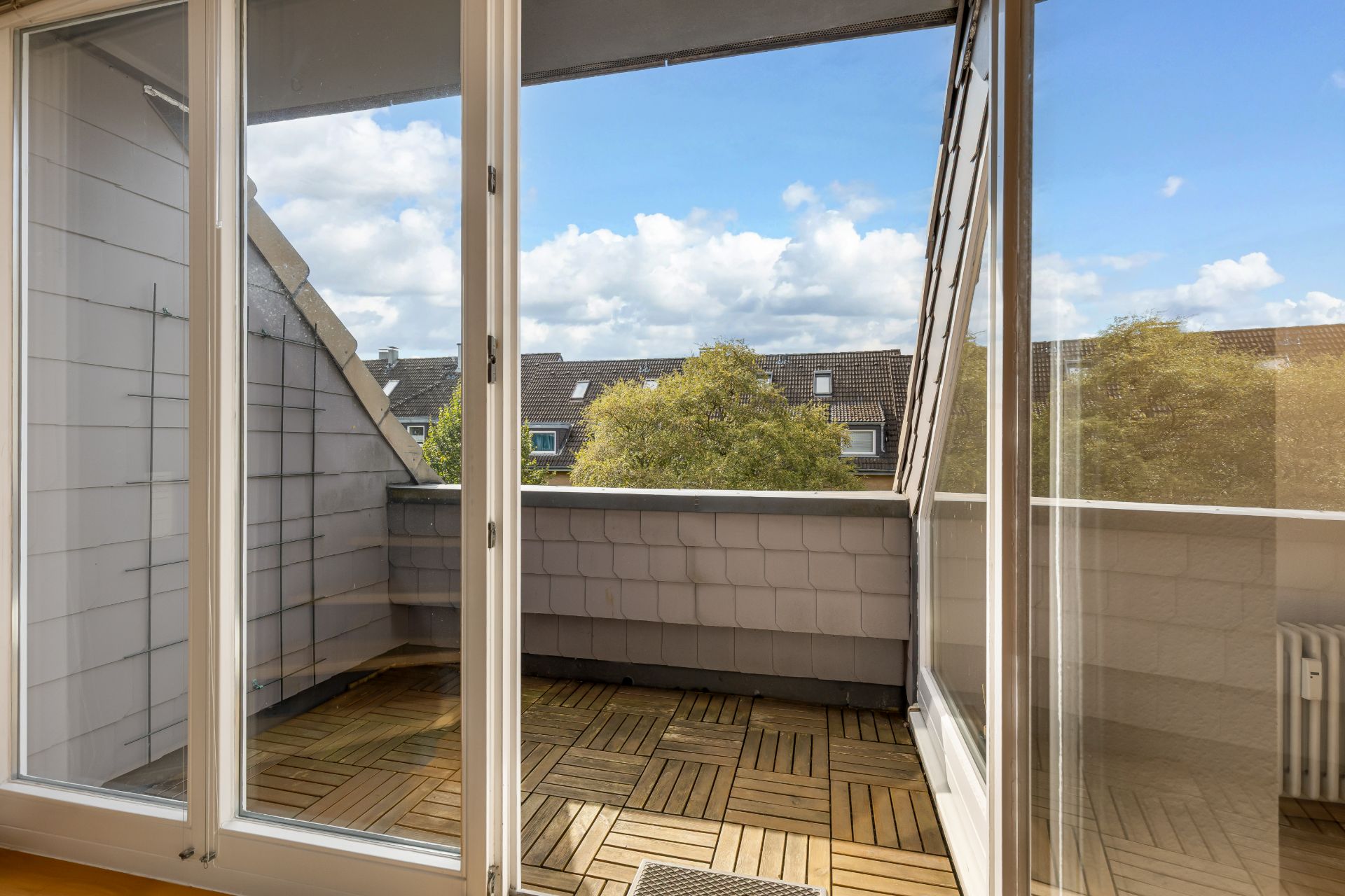 Balkon mit Holzfliesen, Blick durch große Glasfenster auf Bäume und Wohnhäuser.