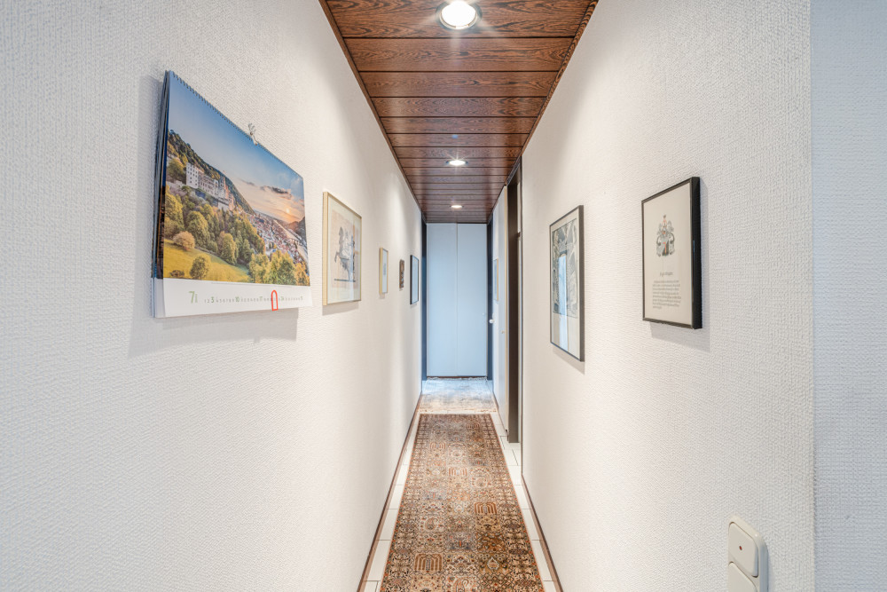 Langer, schmaler Flur mit Teppich, Bildern und Kalender an den Wänden, Holzdecke, helle Beleuchtung.