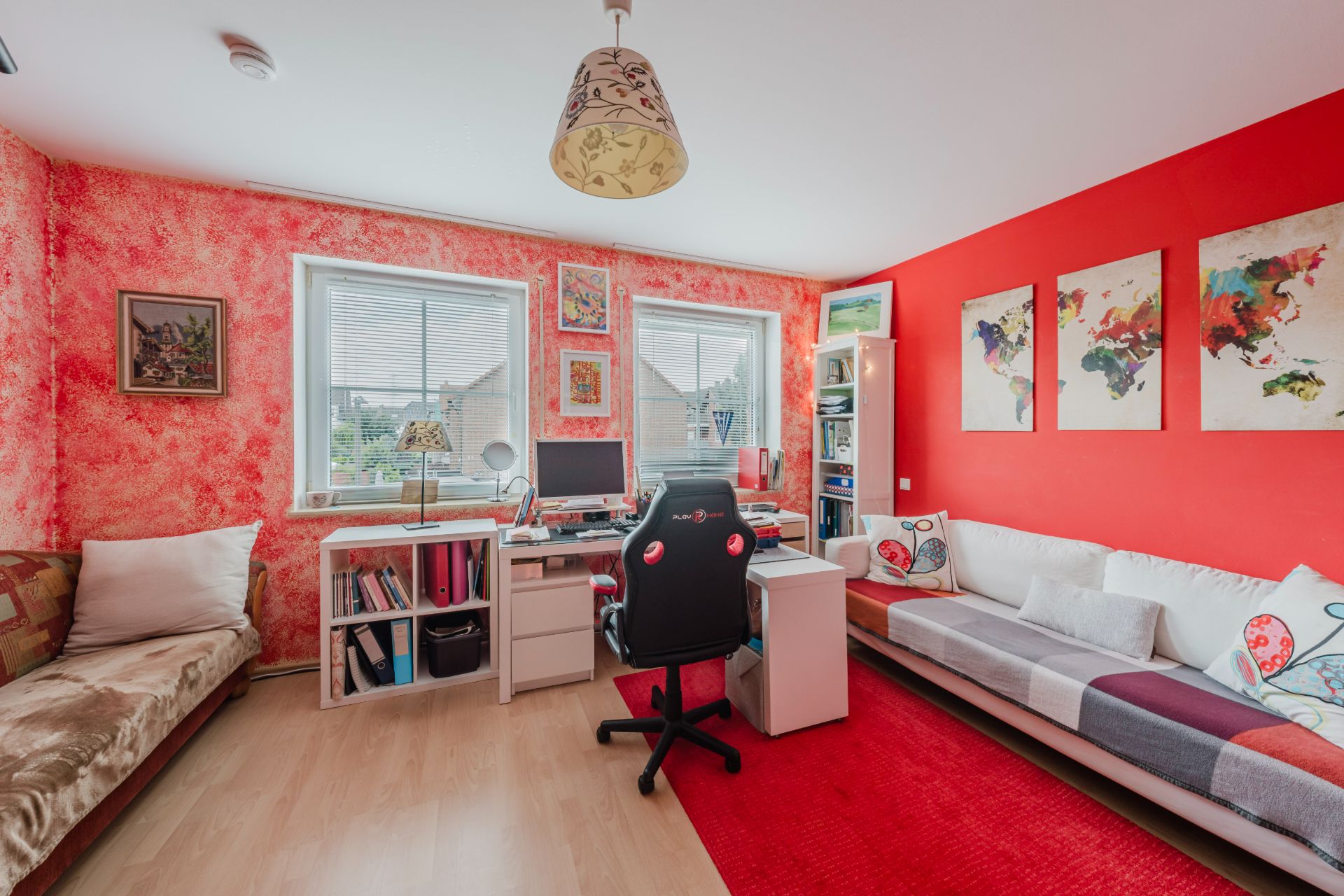 Helles Arbeitszimmer mit zwei Sofas, Schreibtisch, Computer und roter Wand mit bunten Bildern.