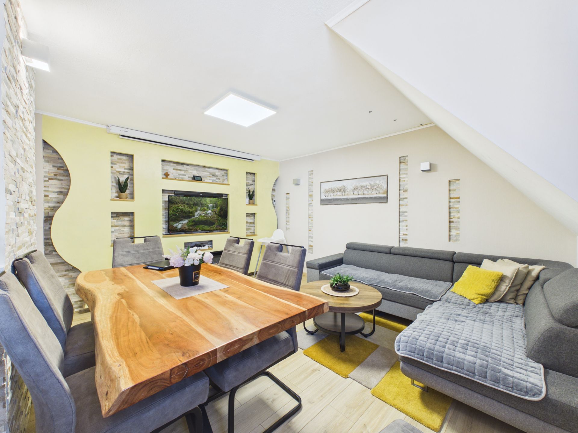 Wohnzimmer mit großem Holztisch, grauen Sofas, TV und gelben Akzenten.