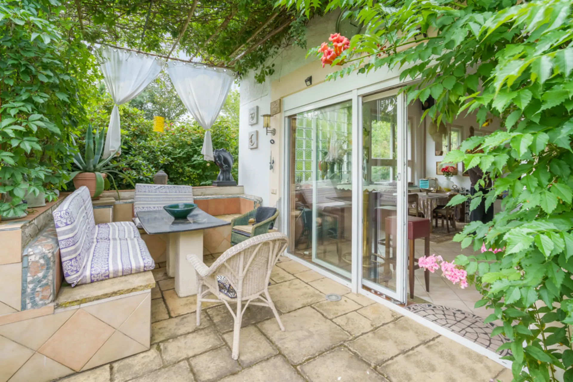 Überdachte Terrasse mit Sitzbank, Tisch und Stühlen neben einer Glastür, umgeben von viel Grün.