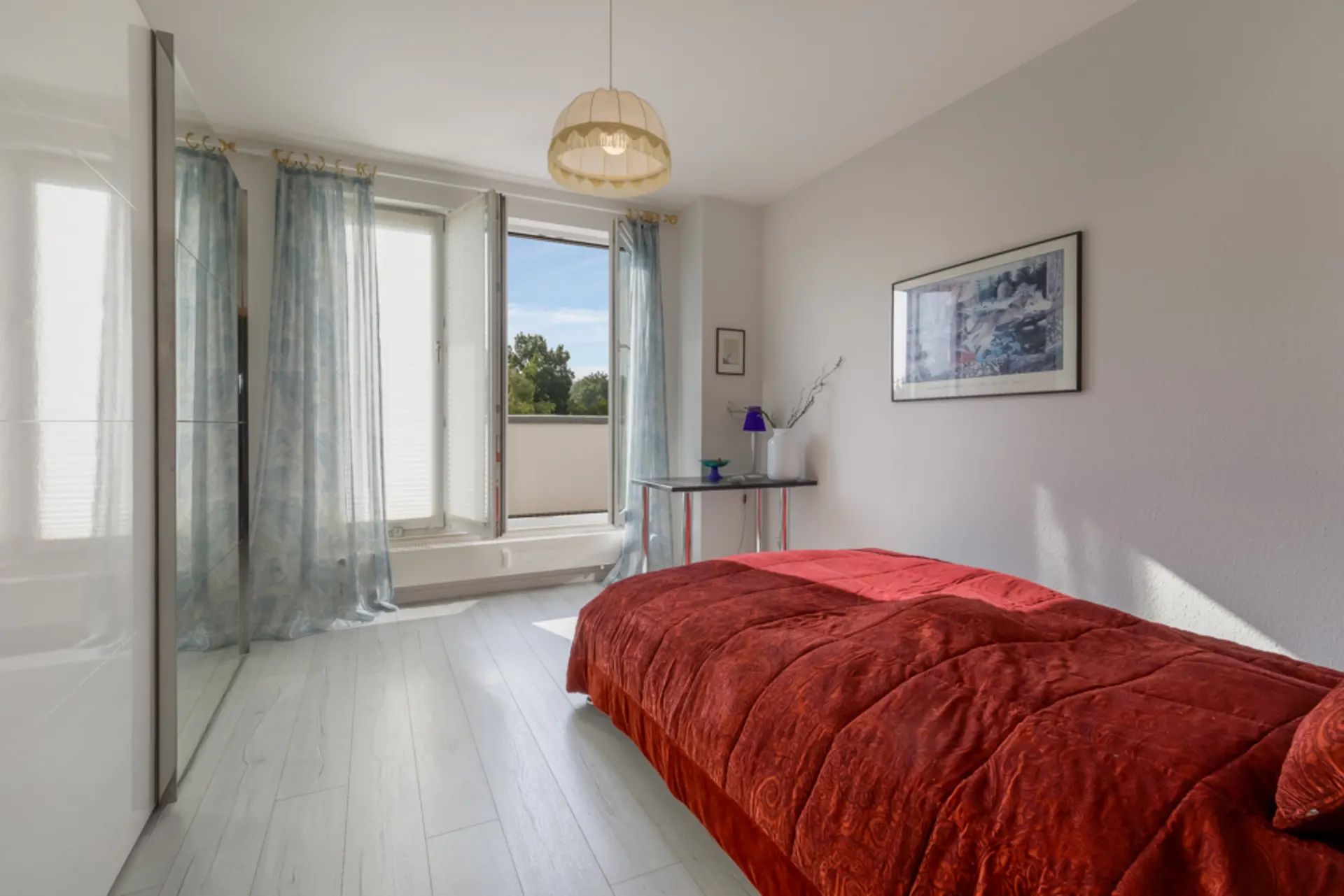 Helles Schlafzimmer mit roter Bettdecke, offenem Fenster, Vorhängen und kleinem Tisch an der Wand.
