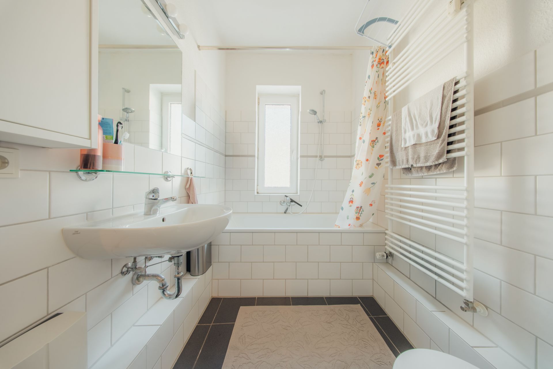 Helles Badezimmer mit Badewanne, Waschbecken, Handtuchheizkörper und Fenster.