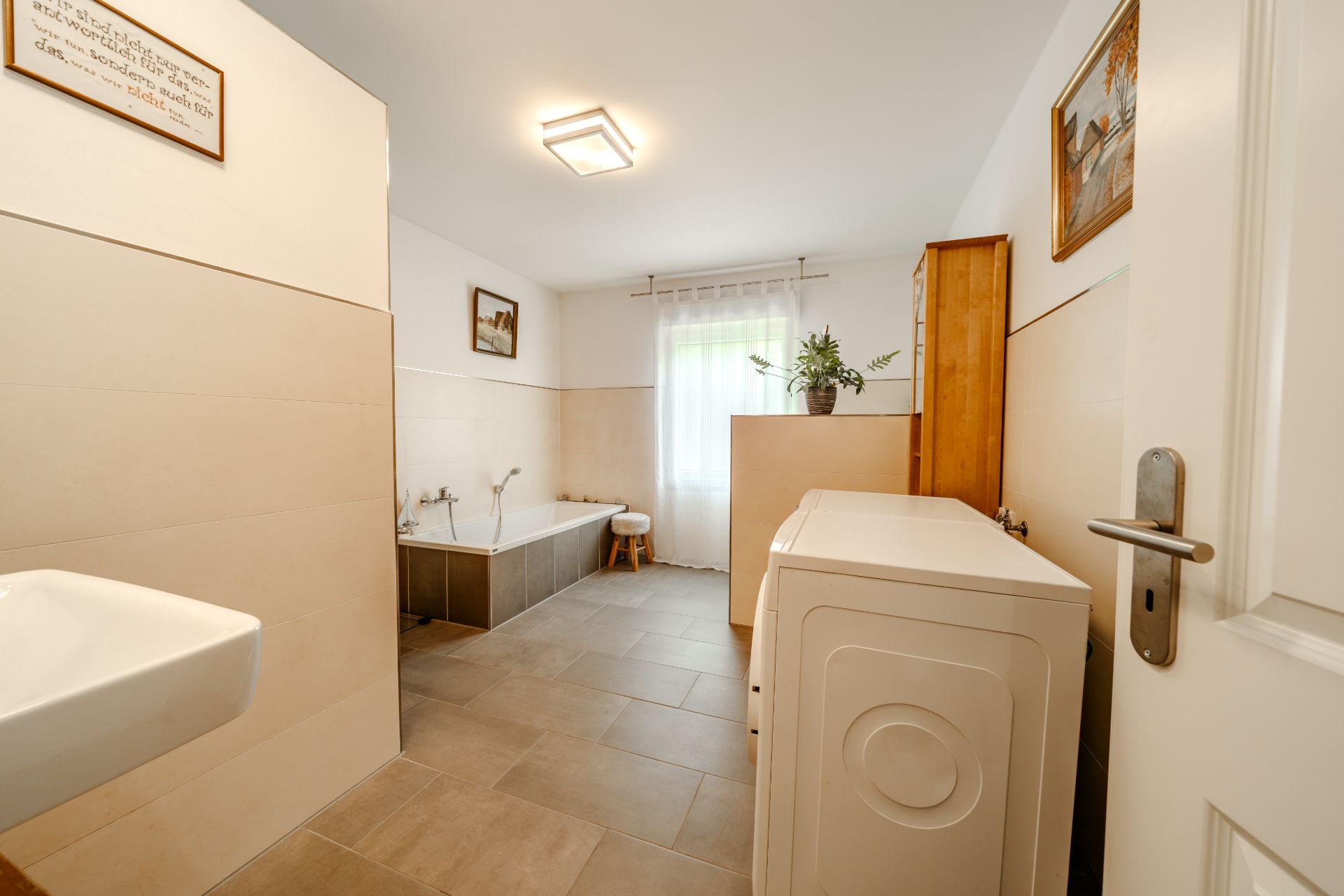 Helles Badezimmer mit Badewanne, Waschmaschine, Waschbecken, Holzschrank und Fenster mit Vorhang.