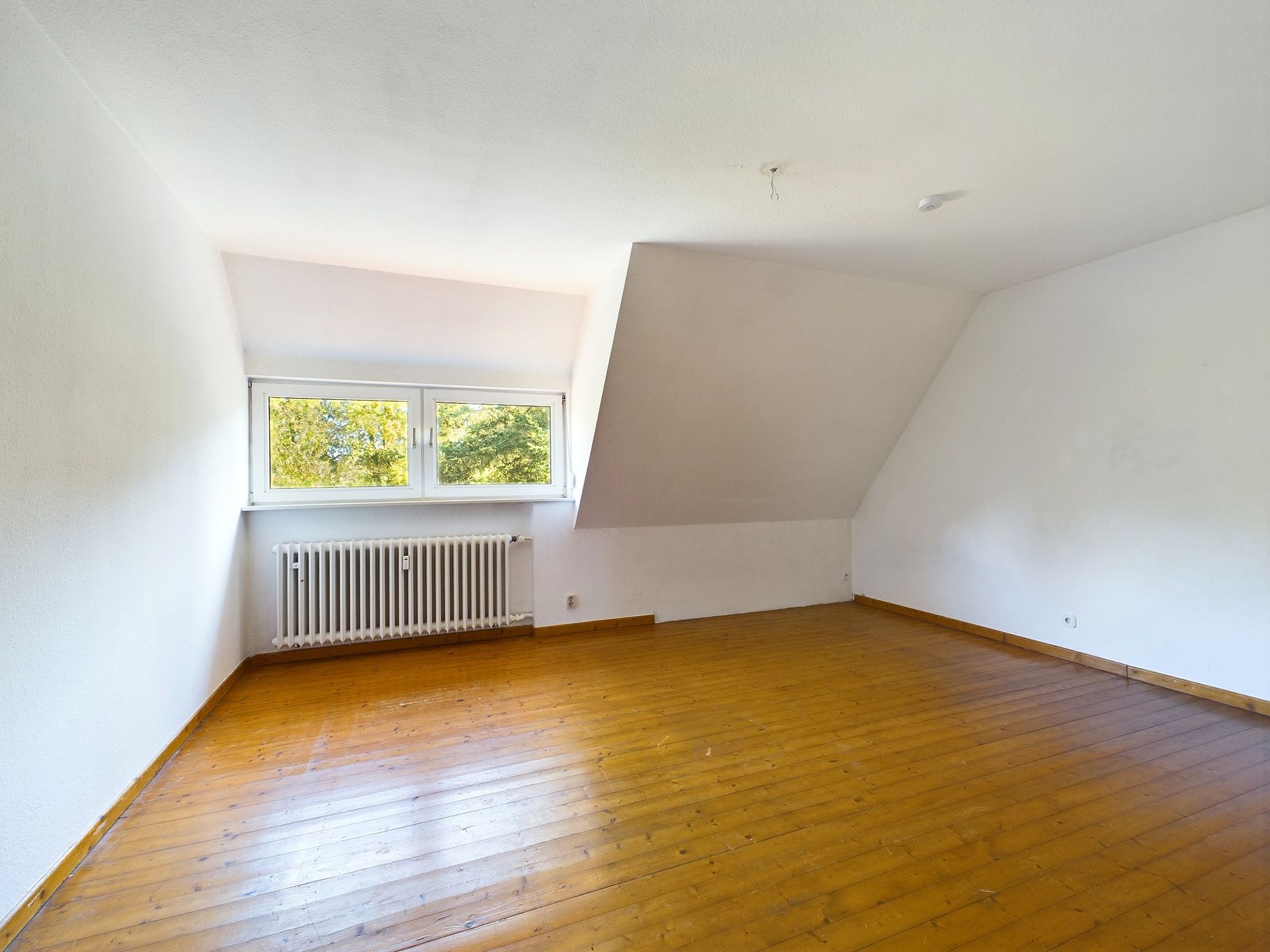 Leeres Zimmer mit schräger Wand, großem Fenster und Holzfußboden.
