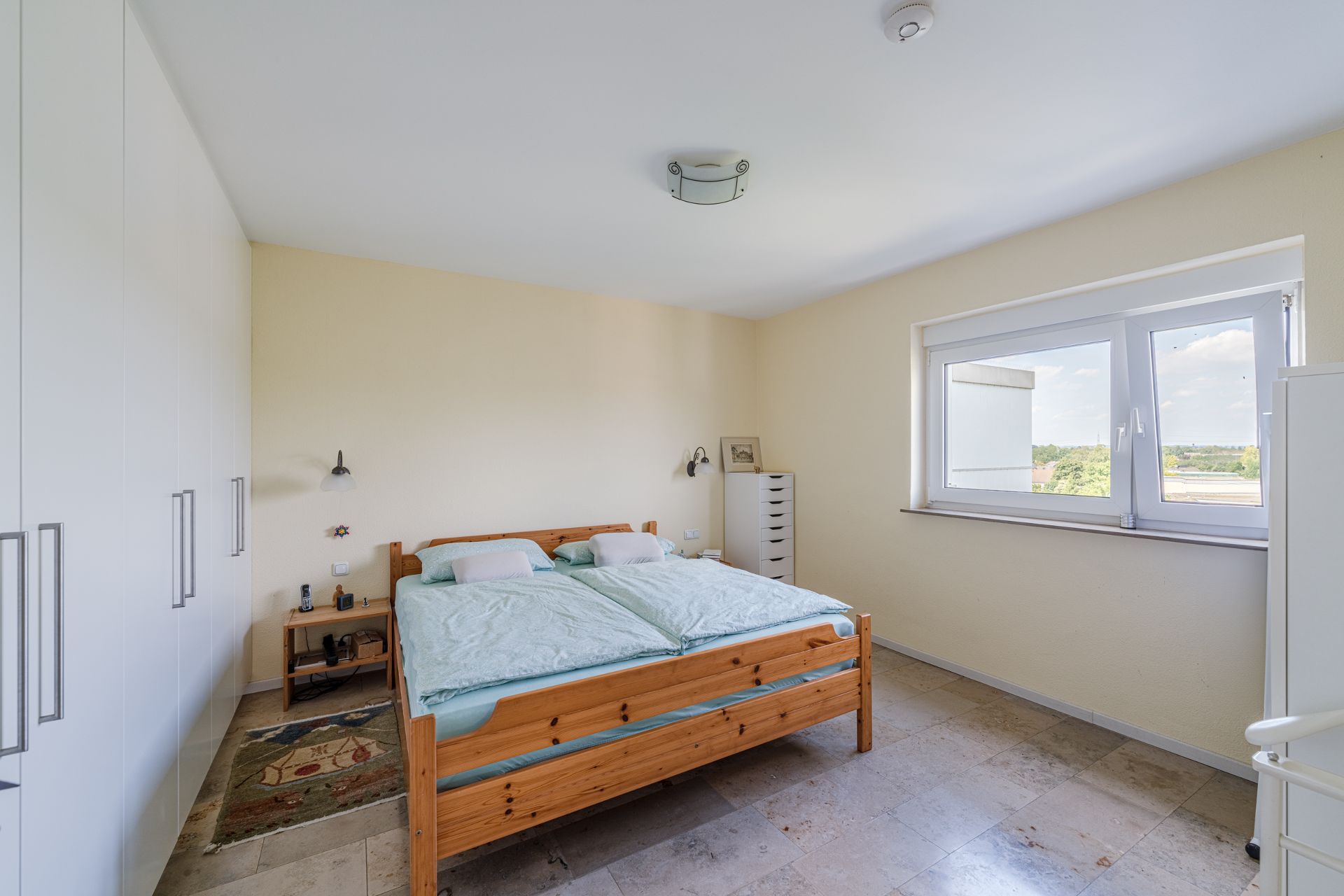 Helles Schlafzimmer mit Holz-Doppelbett, großem Fenster und weißen Einbauschränken.
