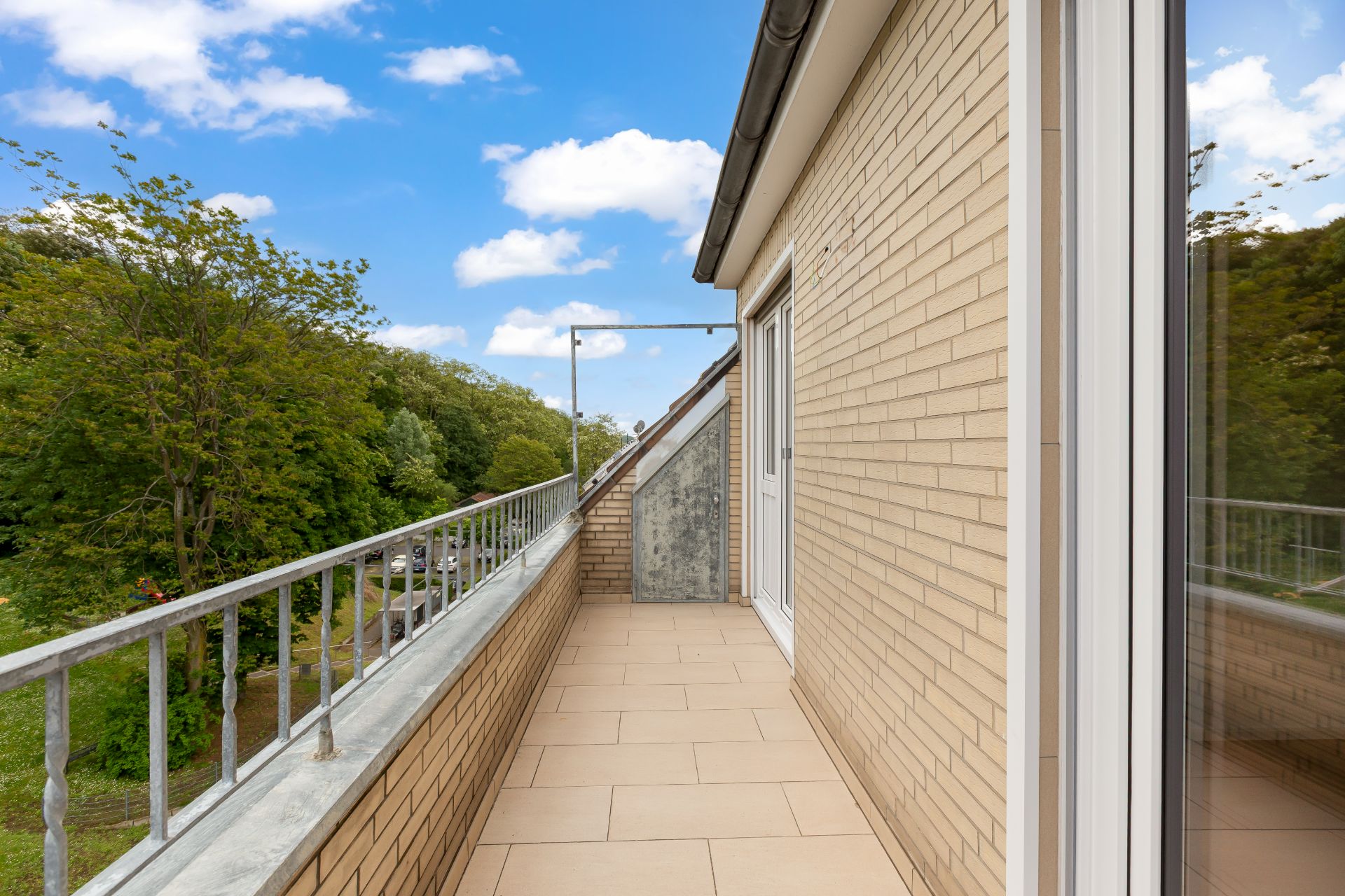 Langer Balkon mit Fliesenboden an einem Wohnhaus, mit Blick auf Bäume und blauem Himmel.