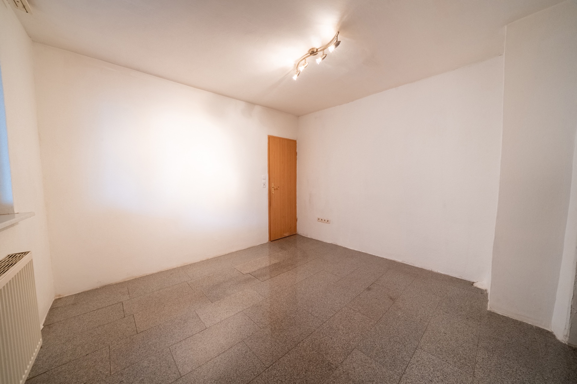 Leerer Raum mit weißen Wänden, grauem Boden, Holztür und Deckenbeleuchtung.