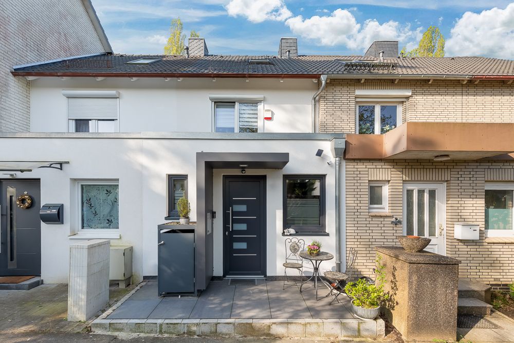 Eingang eines Reihenhauses mit kleiner Terrasse, Tisch, zwei Stühlen und Blumenkübeln.