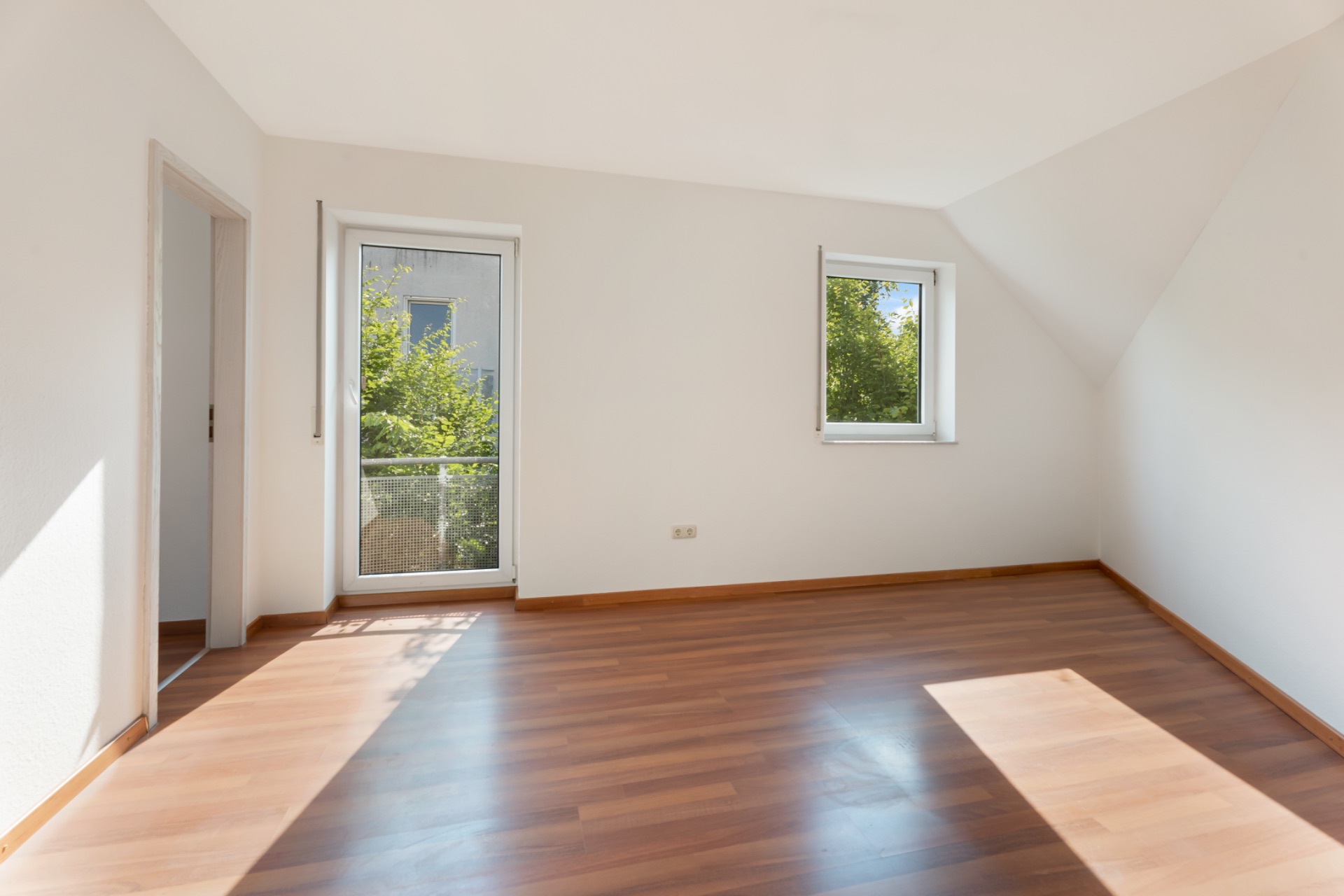 Helles, leeres Zimmer mit Holzfußboden, Fenster und Glastür zu einem kleinen Balkon.