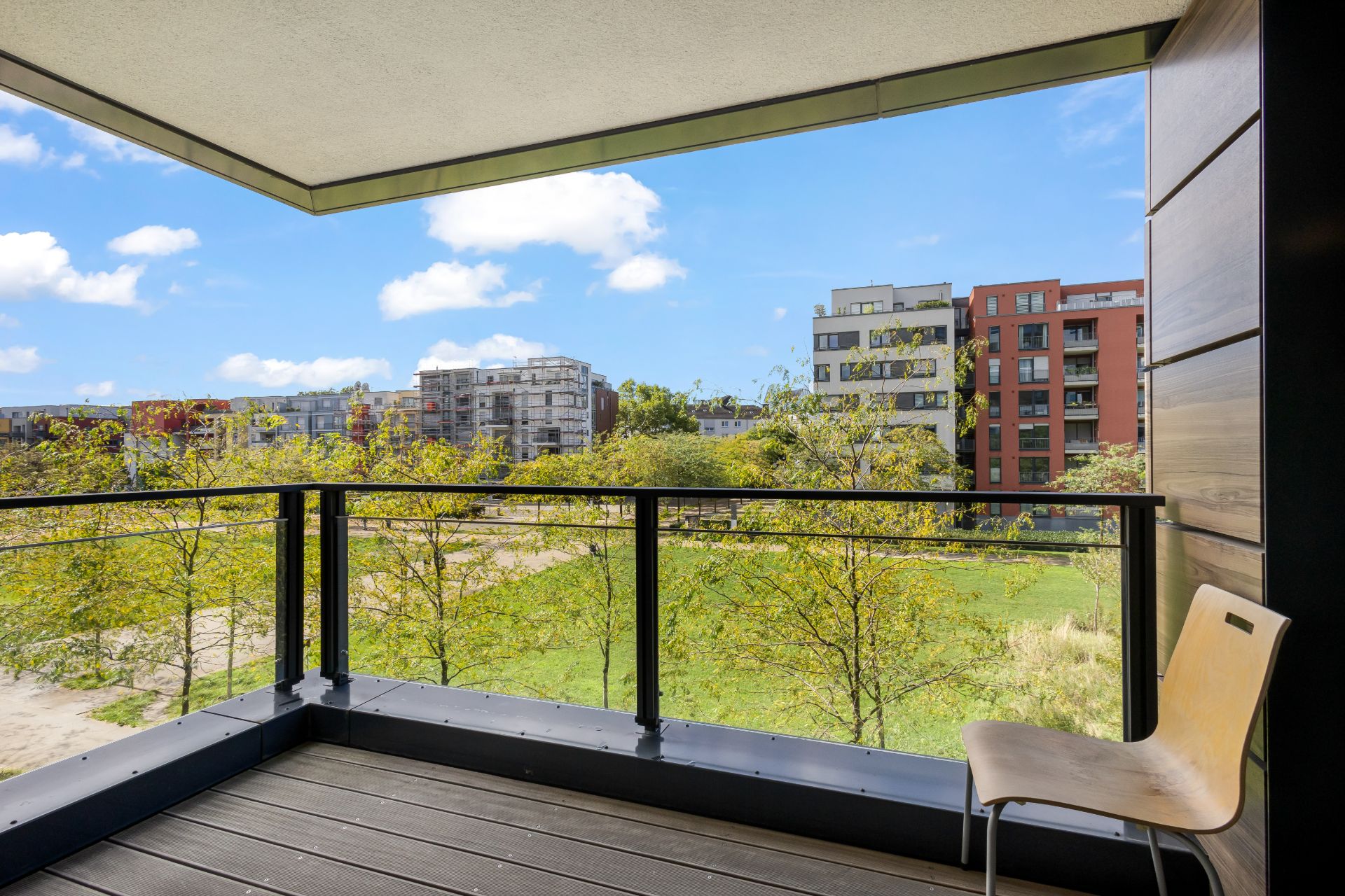 Balkon mit Glasgeländer, Stuhl und Blick auf grüne Bäume und Wohngebäude.