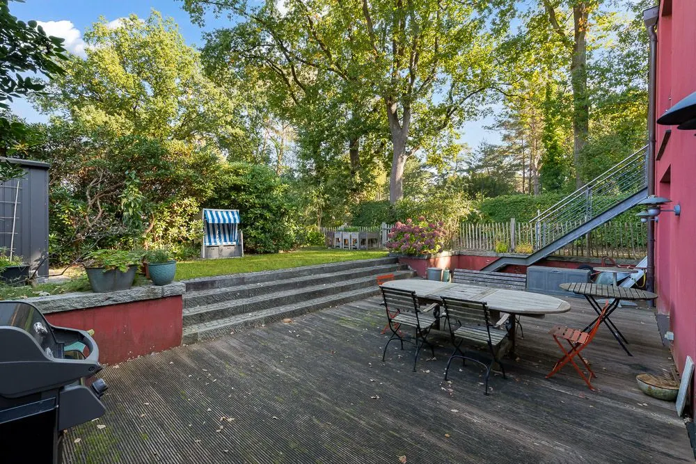 Holzterrasse mit Tisch und Stühlen, Stufen zur Wiese, Bäume und Garten im Hintergrund.