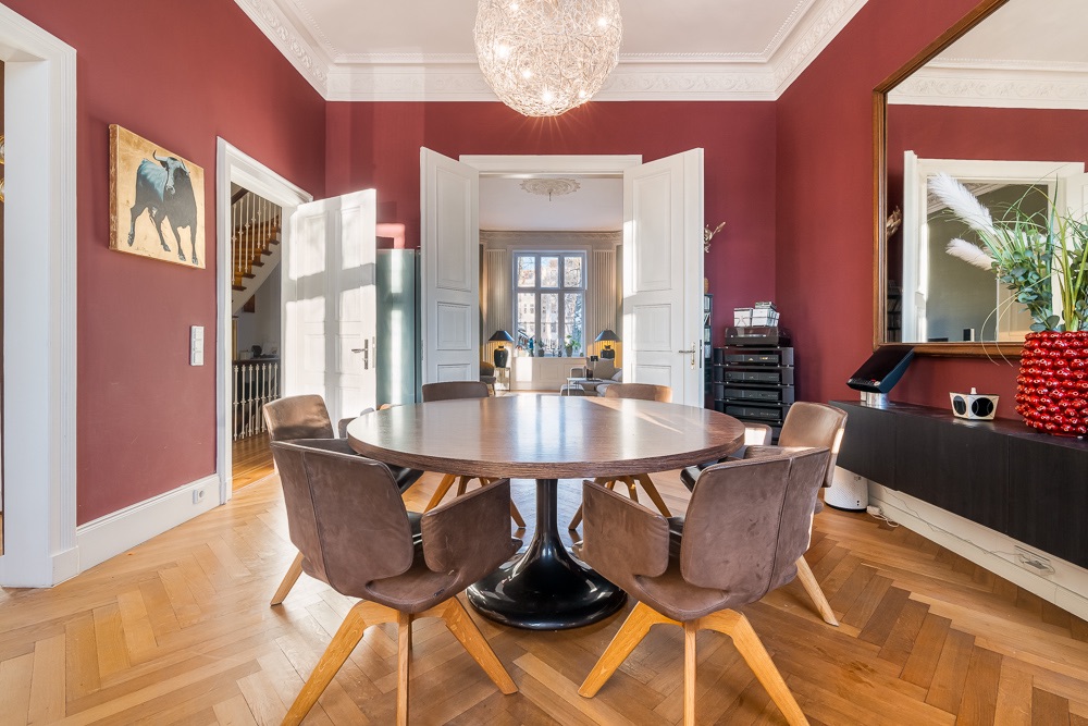 Esszimmer mit rundem Holztisch, sechs Stühlen, roter Wand und großem Spiegel.