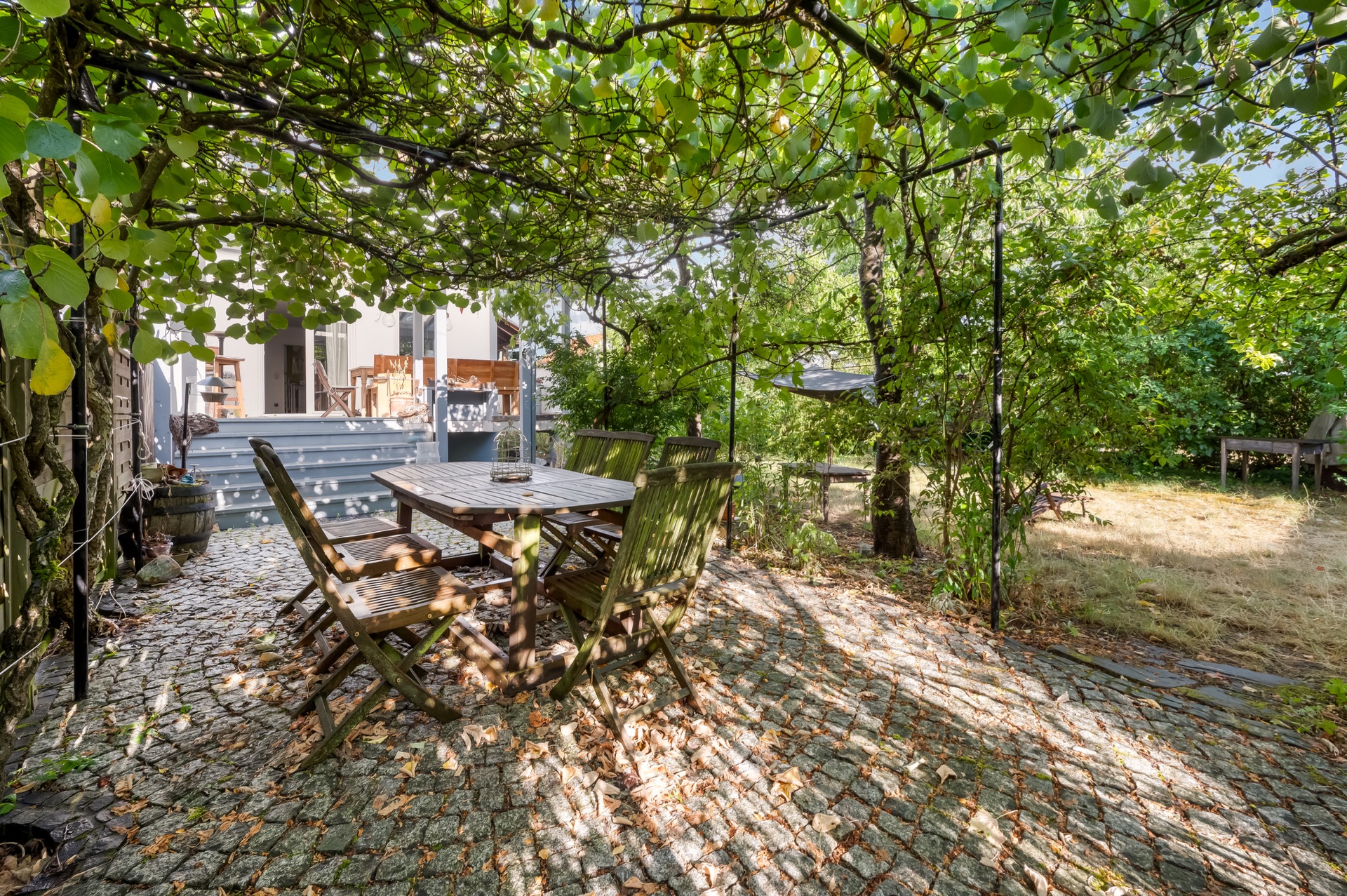 Terrasse mit Holztisch und Stühlen unter einer Laube, umgeben von viel Grün.