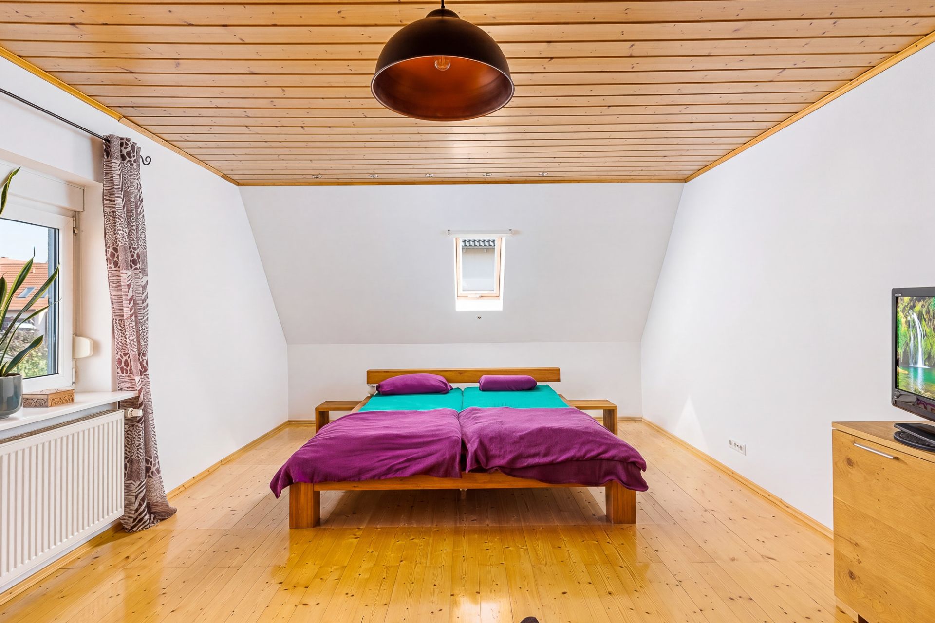 Schlafzimmer mit Holzbett, bunter Bettwäsche, kleinem Fenster und Fernseher auf Holzkommode.