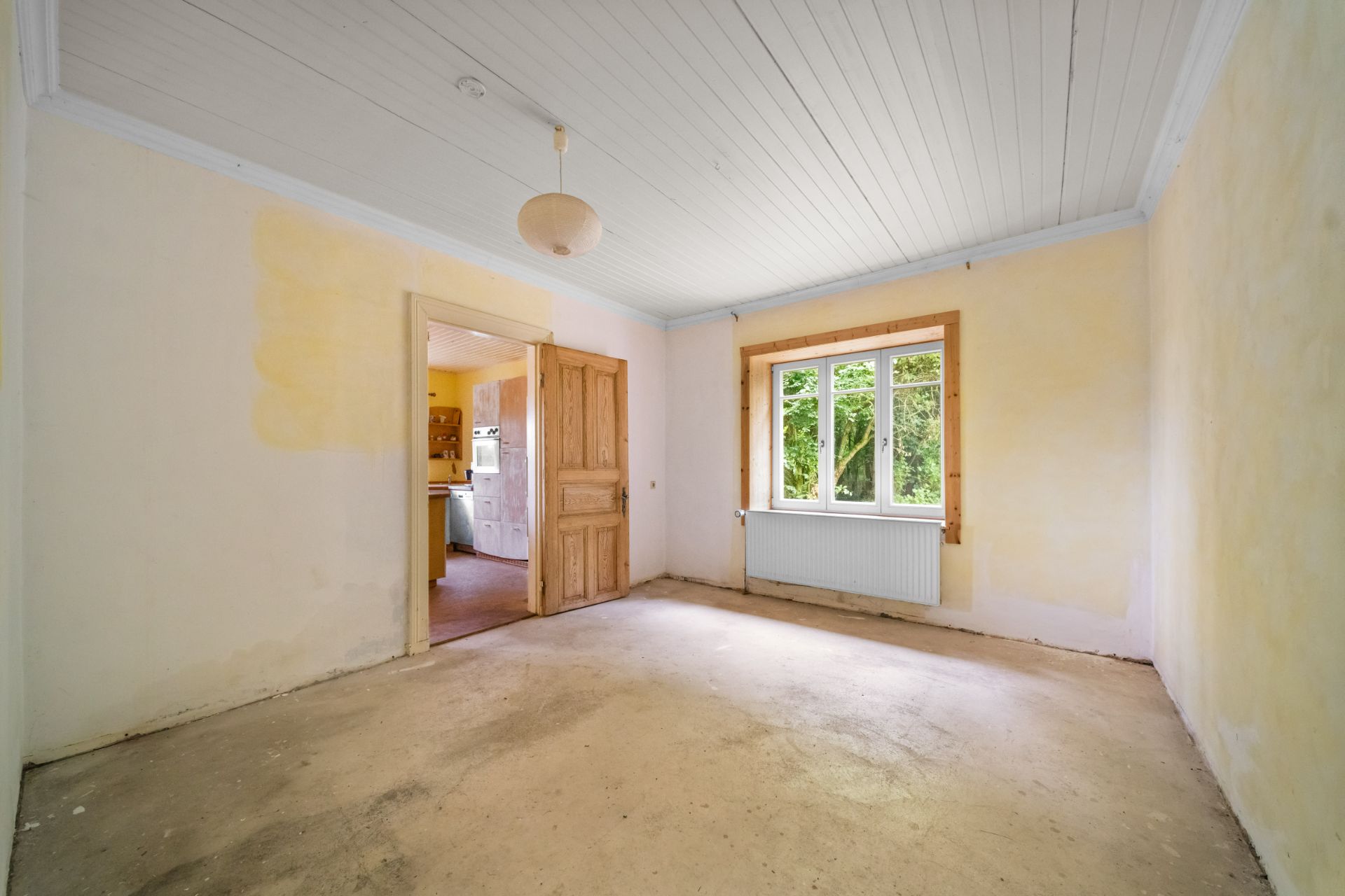Leerer Raum mit Holztür, Fenster, Heizung und sichtbarem Eingangsbereich zur Küche.