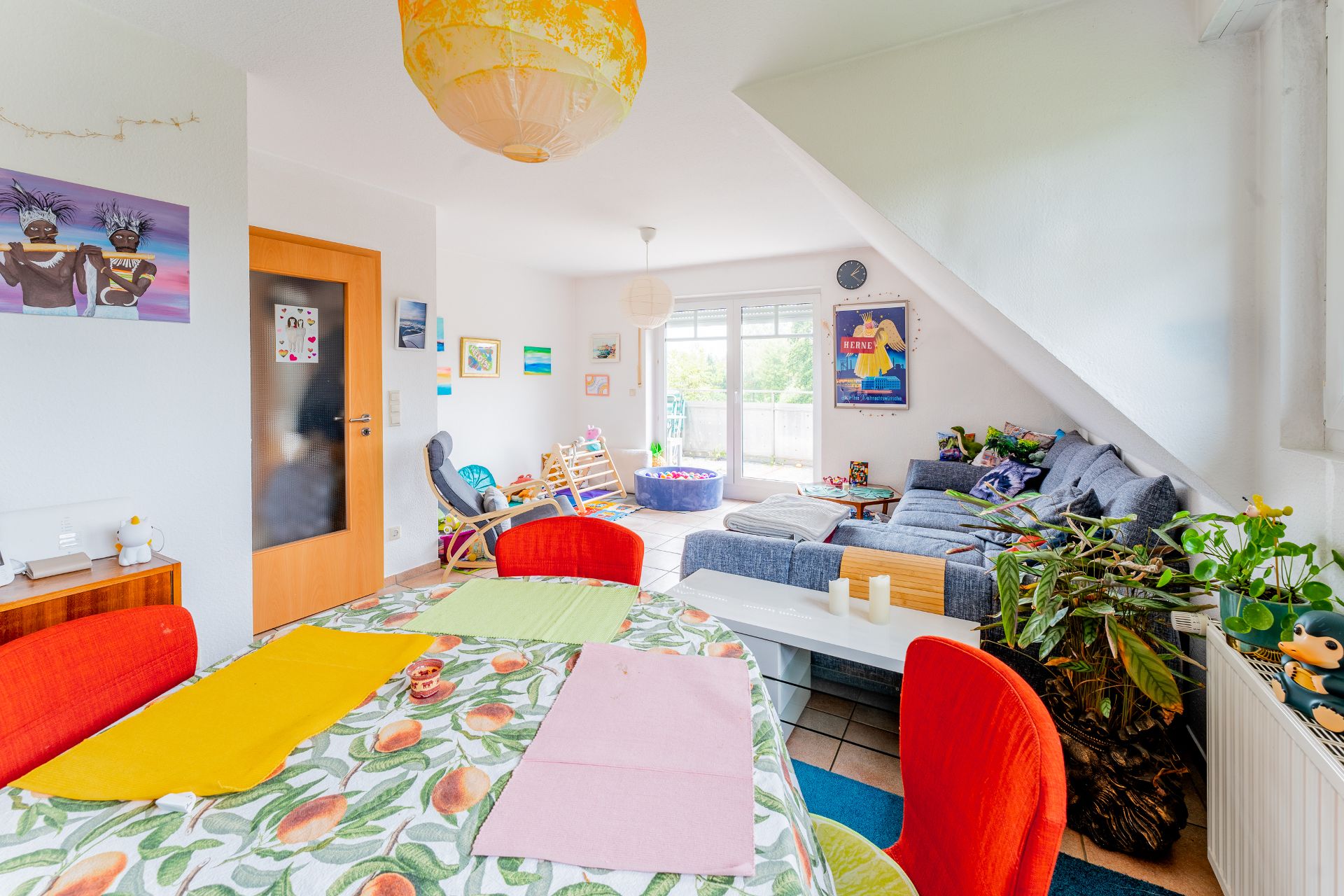 Helles Wohnzimmer mit Esstisch, bunter Dekoration, grauem Sofa und vielen Pflanzen.