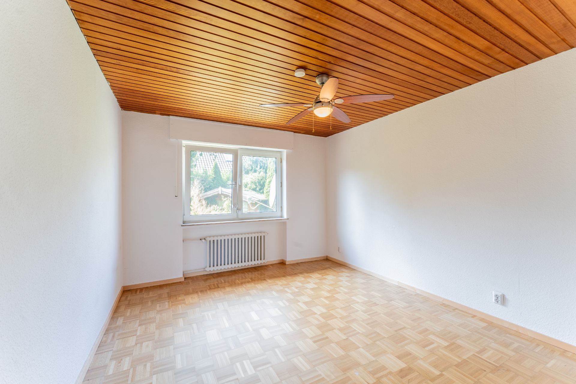 Leeres Zimmer mit Parkettboden, Holzdecke und Deckenventilator, Fenster mit Blick nach draußen.