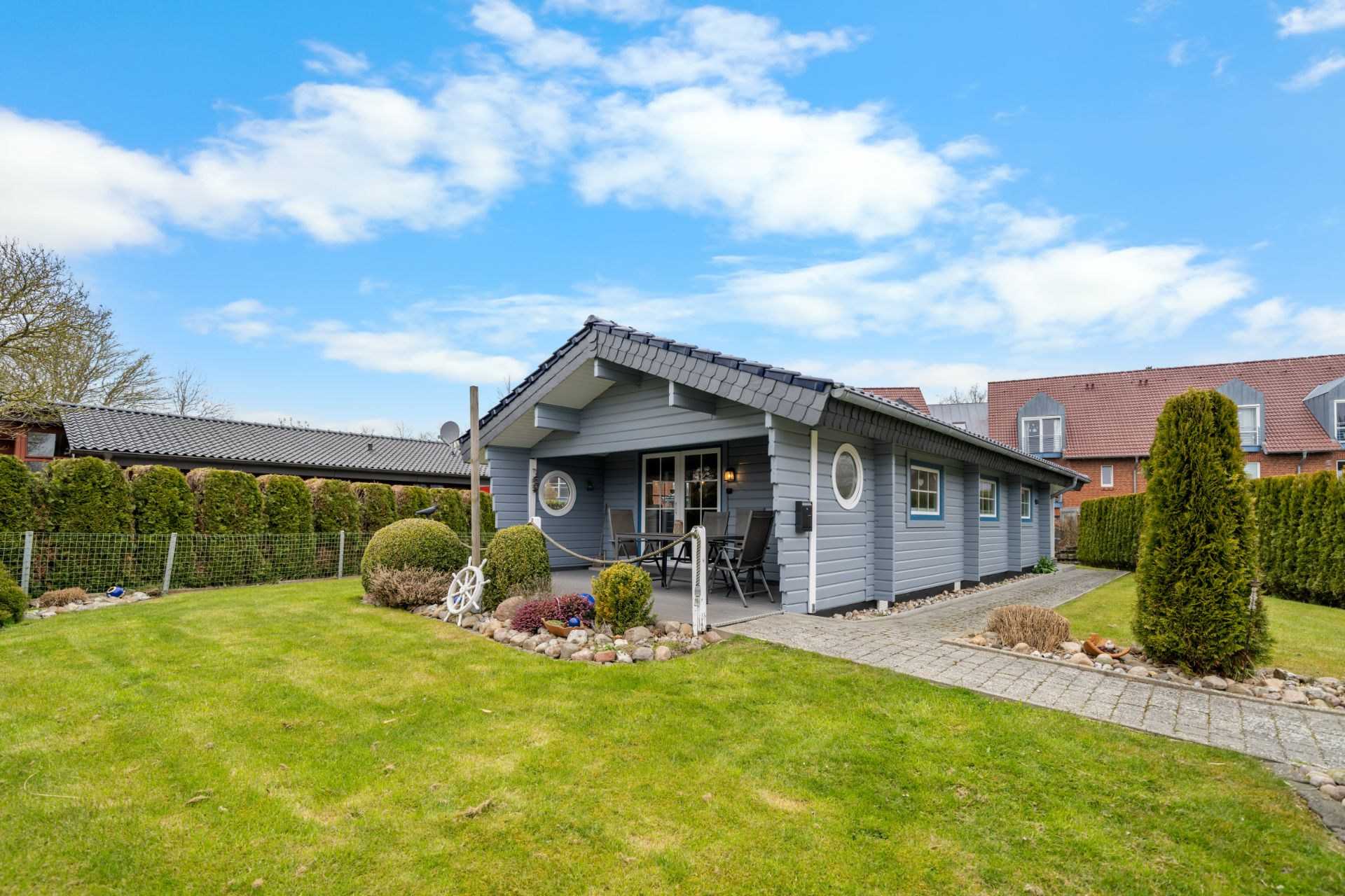Ein einstöckiges graues Haus mit Veranda, Garten und gepflastertem Weg, umgeben von Hecken.