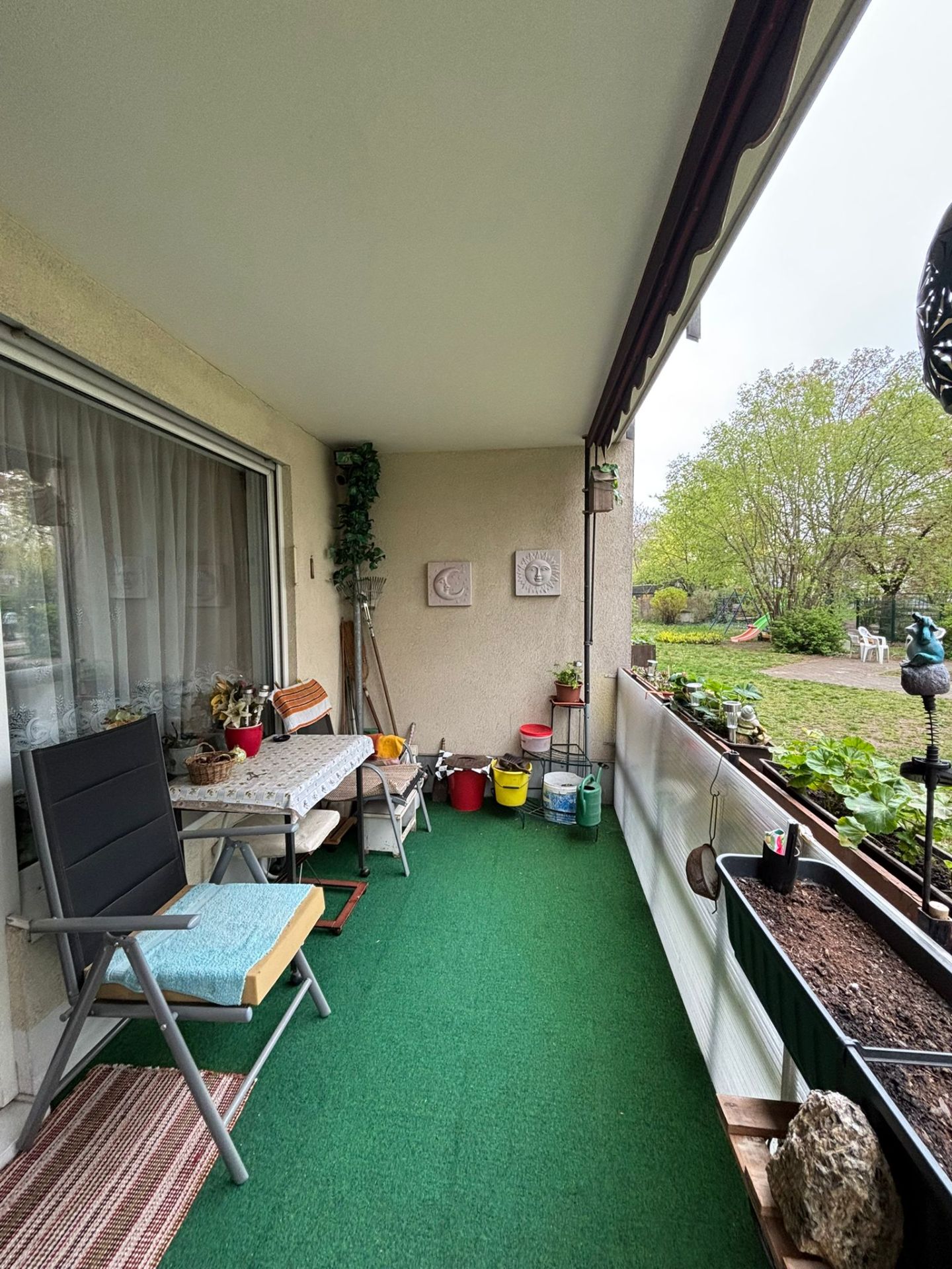Überdachter Balkon mit grünem Teppich, Stuhl, Tisch, Pflanzen und Blick auf einen Garten.