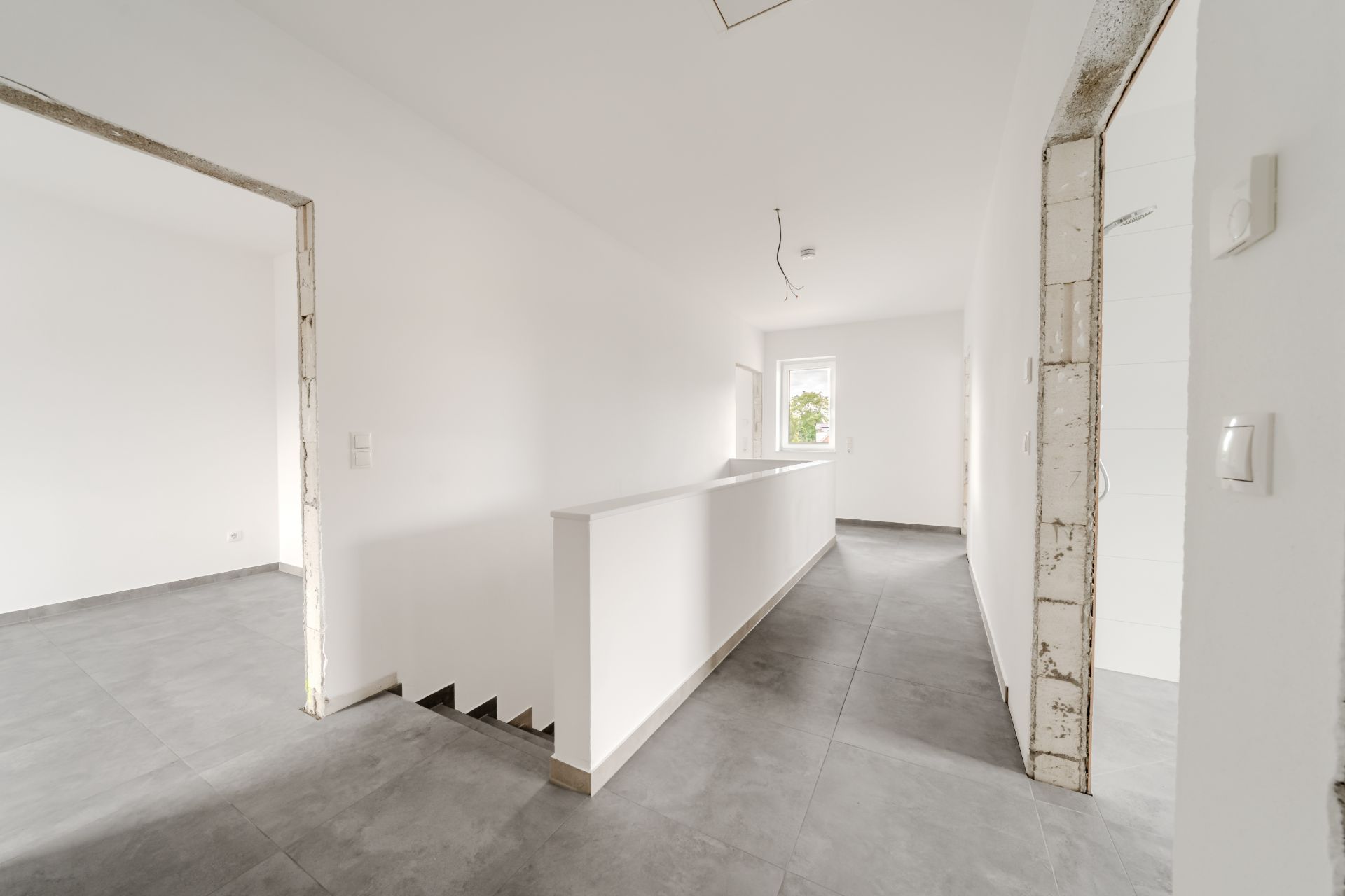 Heller Flur mit grauen Fliesen, offenen Türrahmen und Treppe, Fenster am Ende des Gangs.
