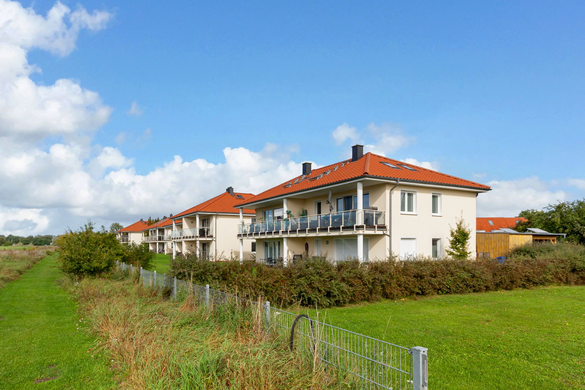 Zwei helle Wohnhäuser mit roten Dächern, Balkonen und viel Grün im Vordergrund.