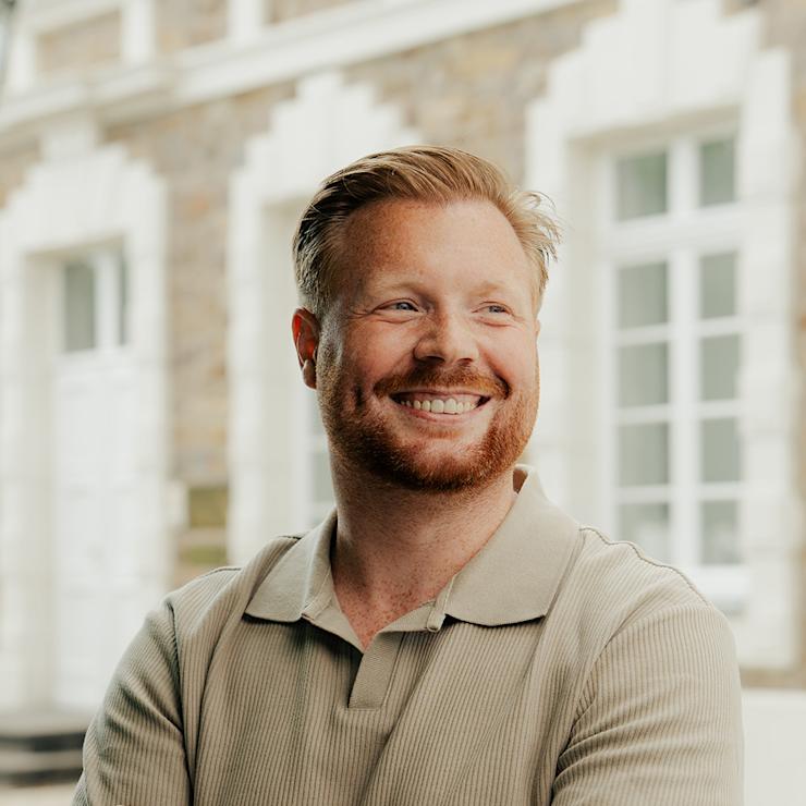 Mann mit bart und tätowiertem Arm steht draußen vor einem Gebäude mit weißen Fenstern.
