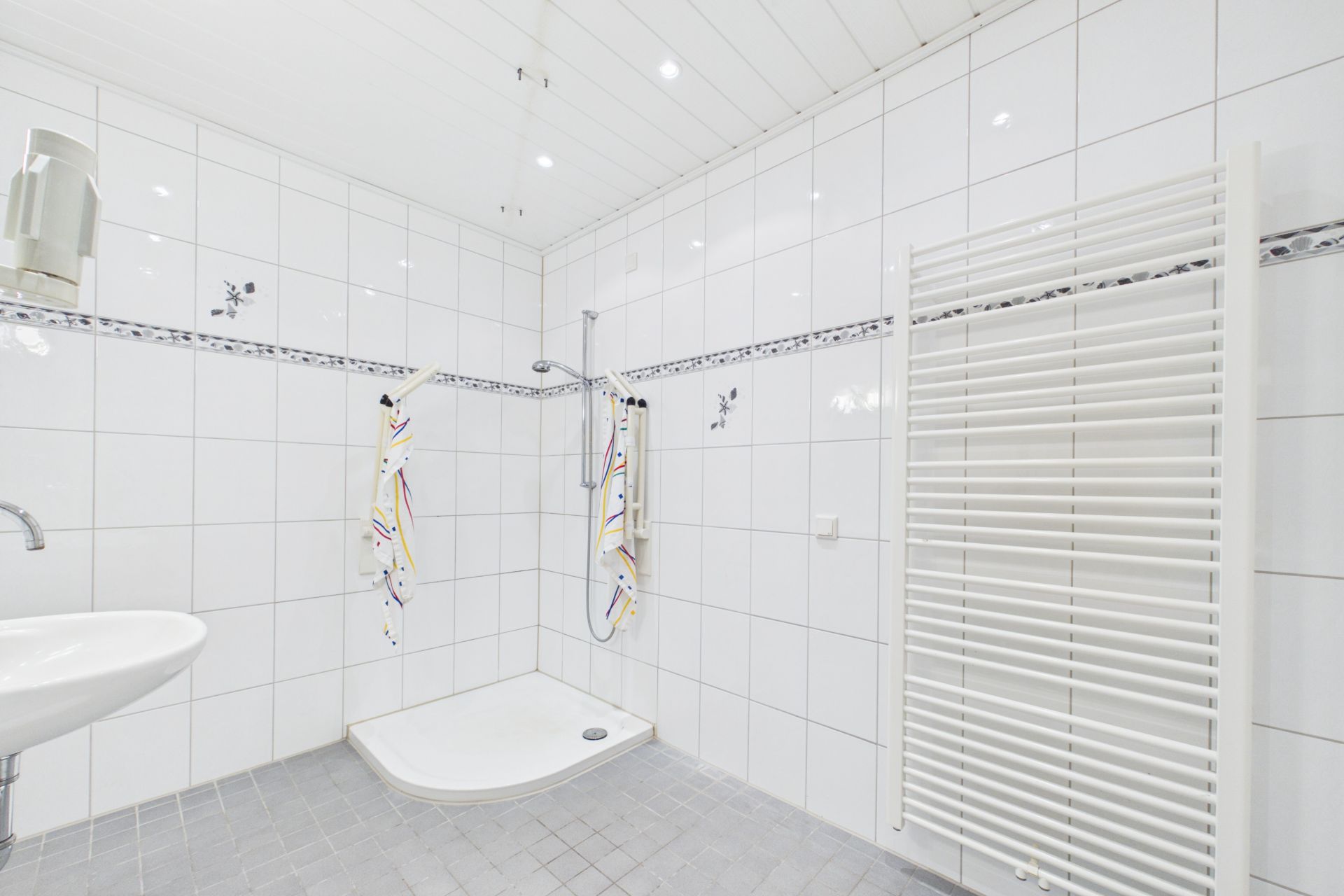 Modernes, weiß gefliestes Bad mit Dusche, Waschbecken, Handtüchern und Heizkörper.