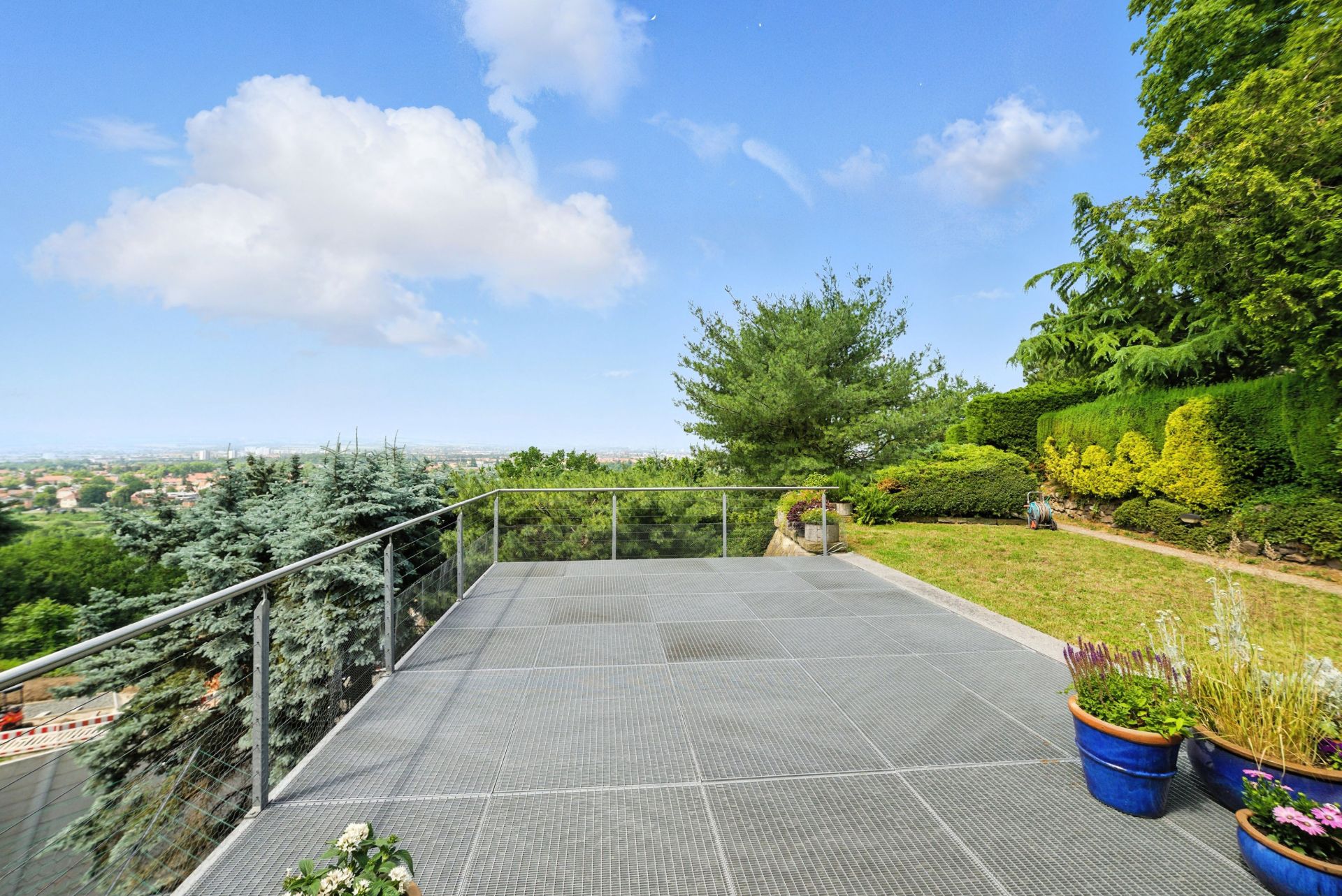 Große Terrasse mit Glasgeländer, Pflanzkübeln und Blick auf Garten und Bäume.