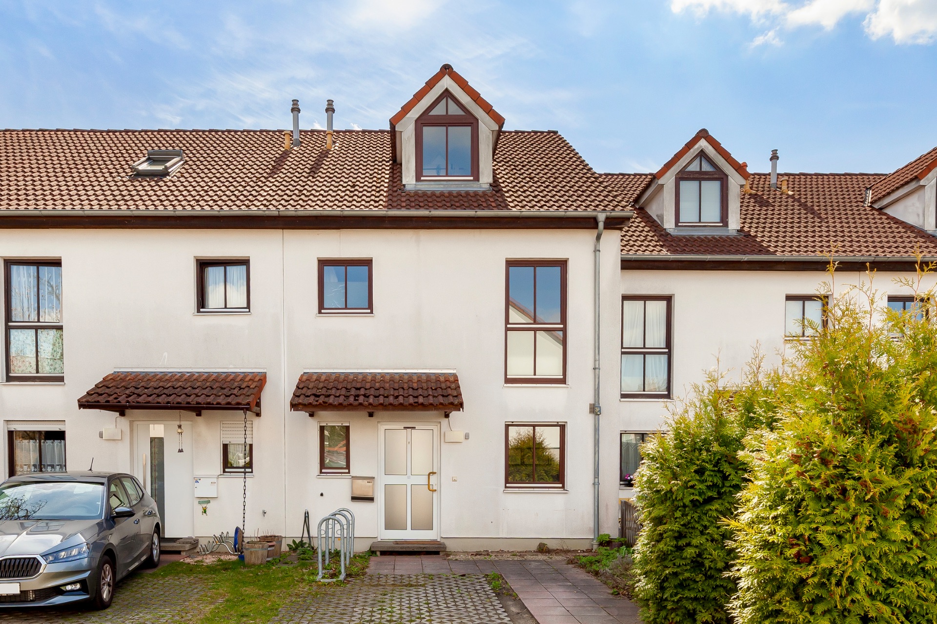Reihenhaus mit roten Dachziegeln, zwei Eingängen und großen Fenstern, davor ein Parkplatz und Sträucher.