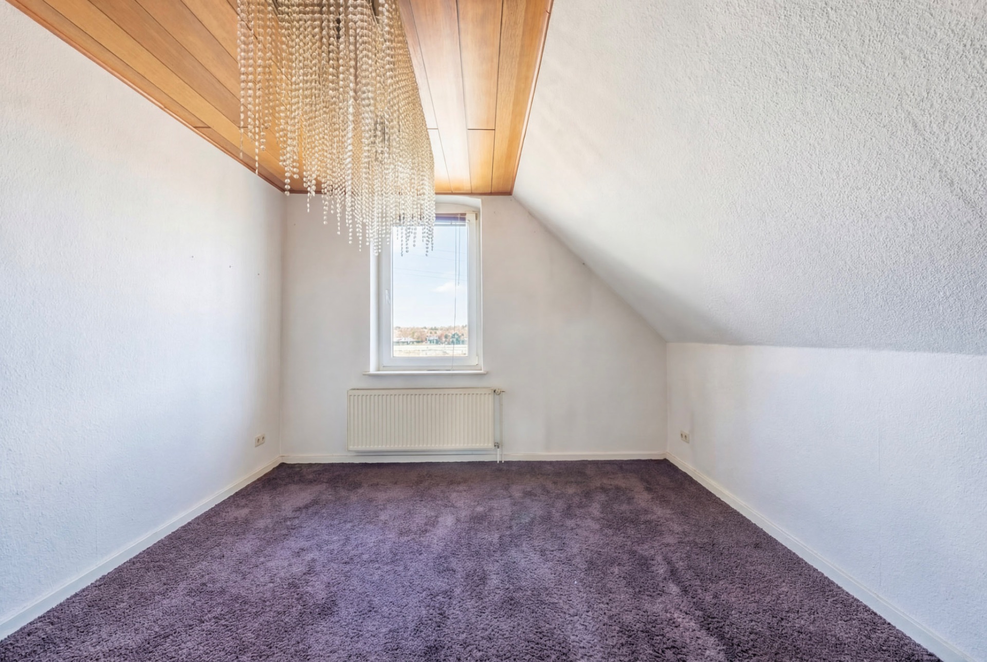 Leeres Zimmer mit lila Teppich, weißer Wand, Holzdecke und Fenster mit Heizkörper.