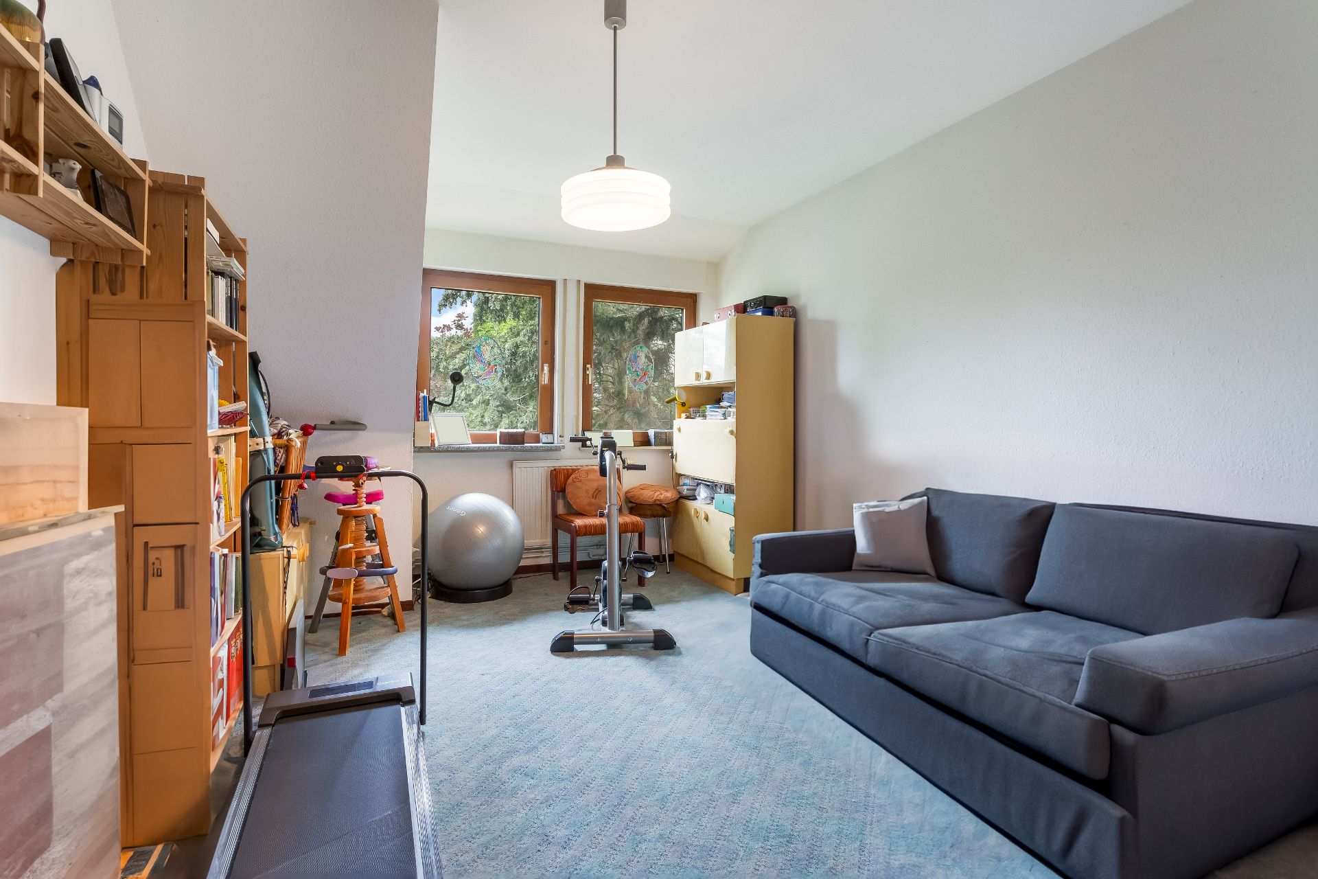 Helles Zimmer mit Sofa, Bücherregal, Fitnessgeräten und zwei Fenstern zum Garten.